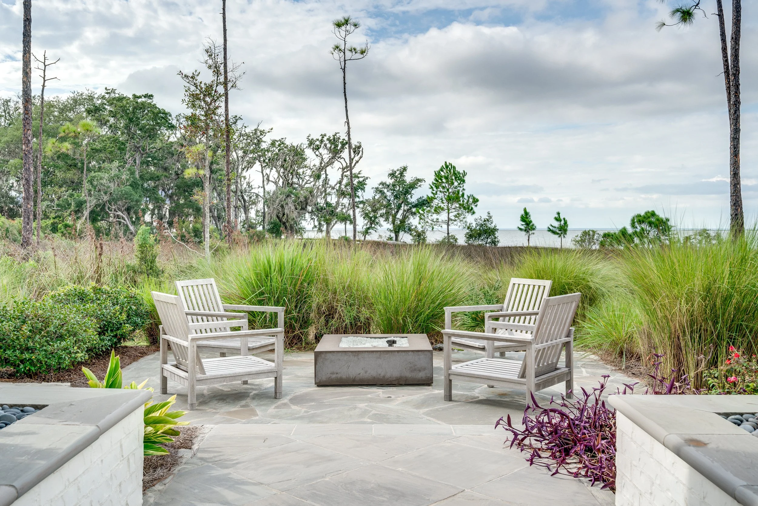 Outdoor patio with two wooden chairs and a fire pit, surrounded by greenery and trees, overlooking a body of water under a partly cloudy sky - Real estate photography in 30A, Destin, and Panama City Beach by Matt Jozwiak