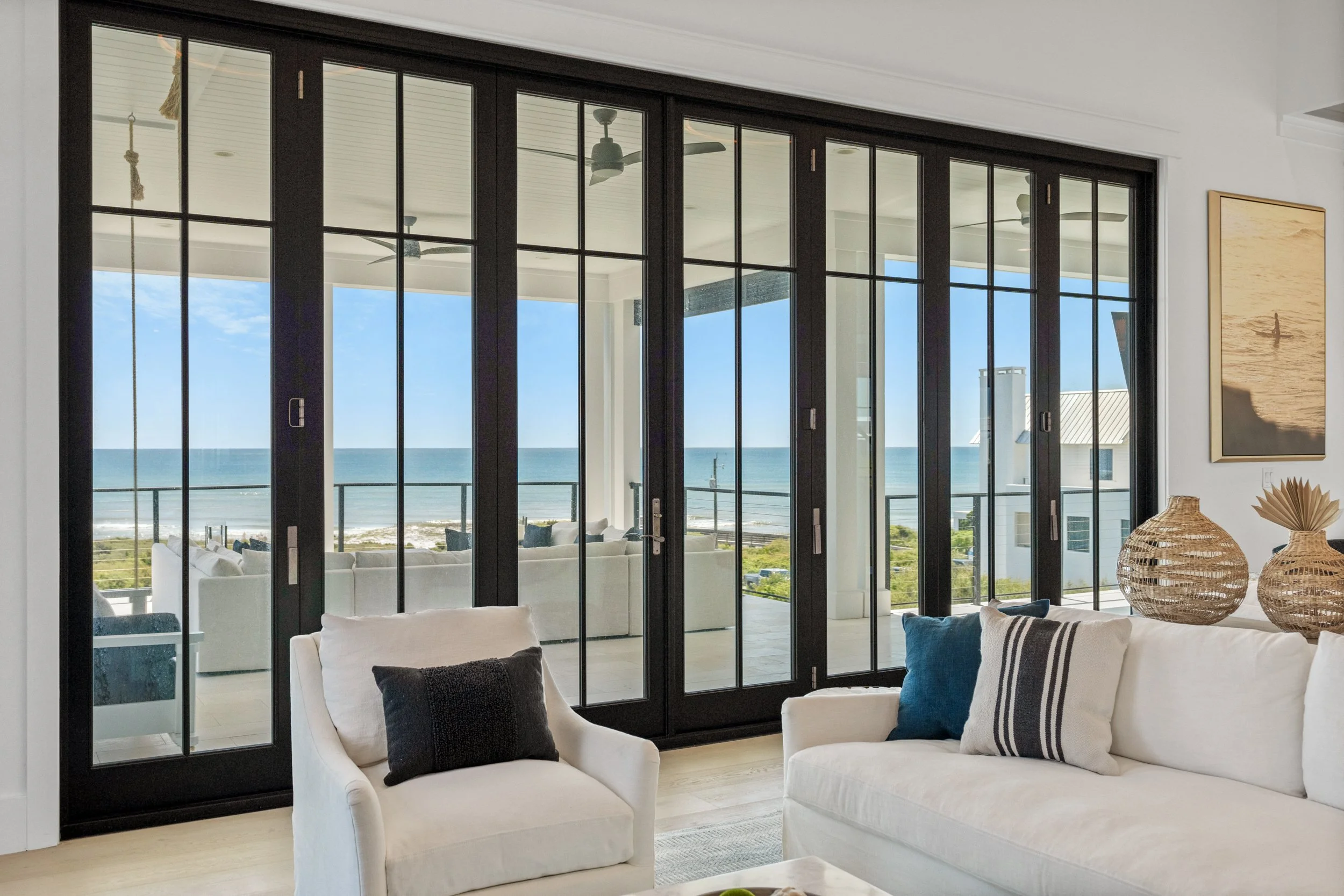 Living room with large black-framed glass sliding doors leading to a balcony overlooking the beach and ocean, with white furniture and decorative pillows - Real estate photography in 30A, Destin, and Panama City Beach by Matt Jozwiak