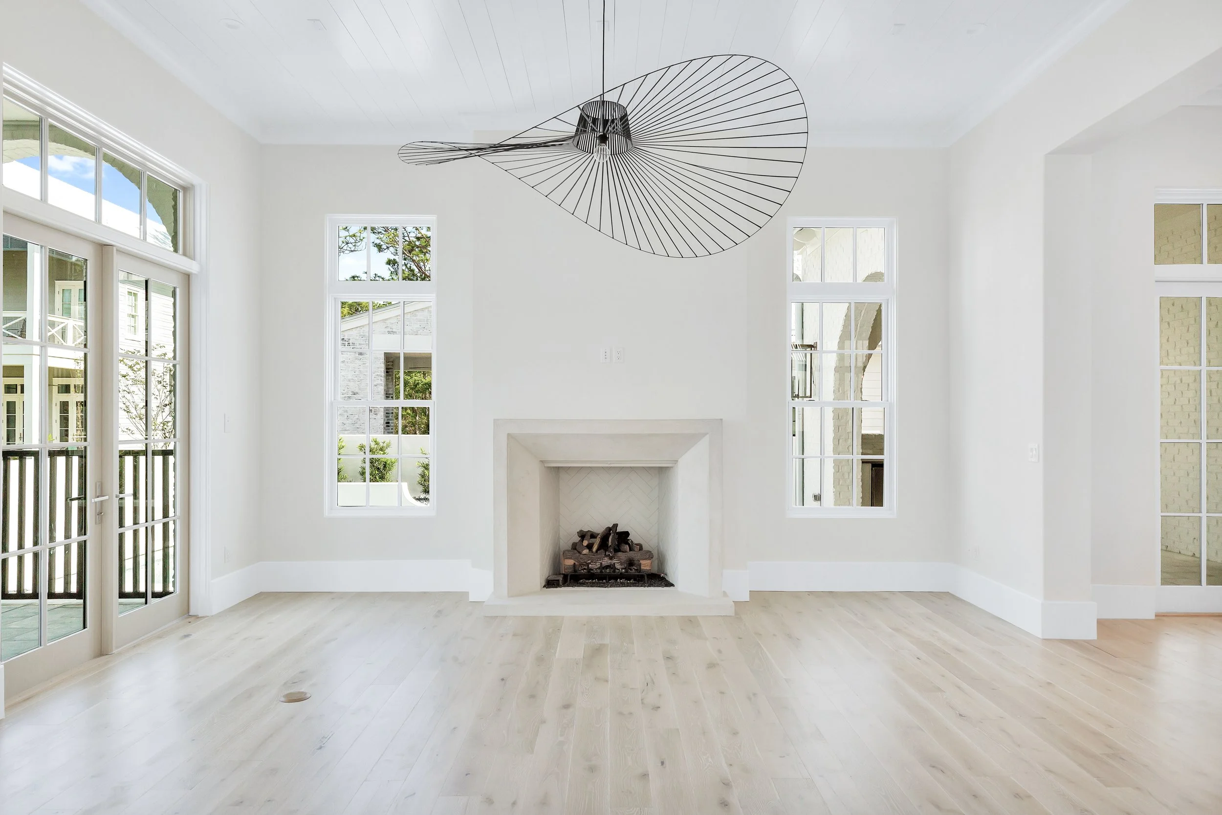 Empty living room with light-colored hardwood floors, white walls, a fireplace, large windows, and a modern black wire pendant light fixture on the ceiling - Real estate photography in 30A, Destin, and Panama City Beach by Matt Jozwiak