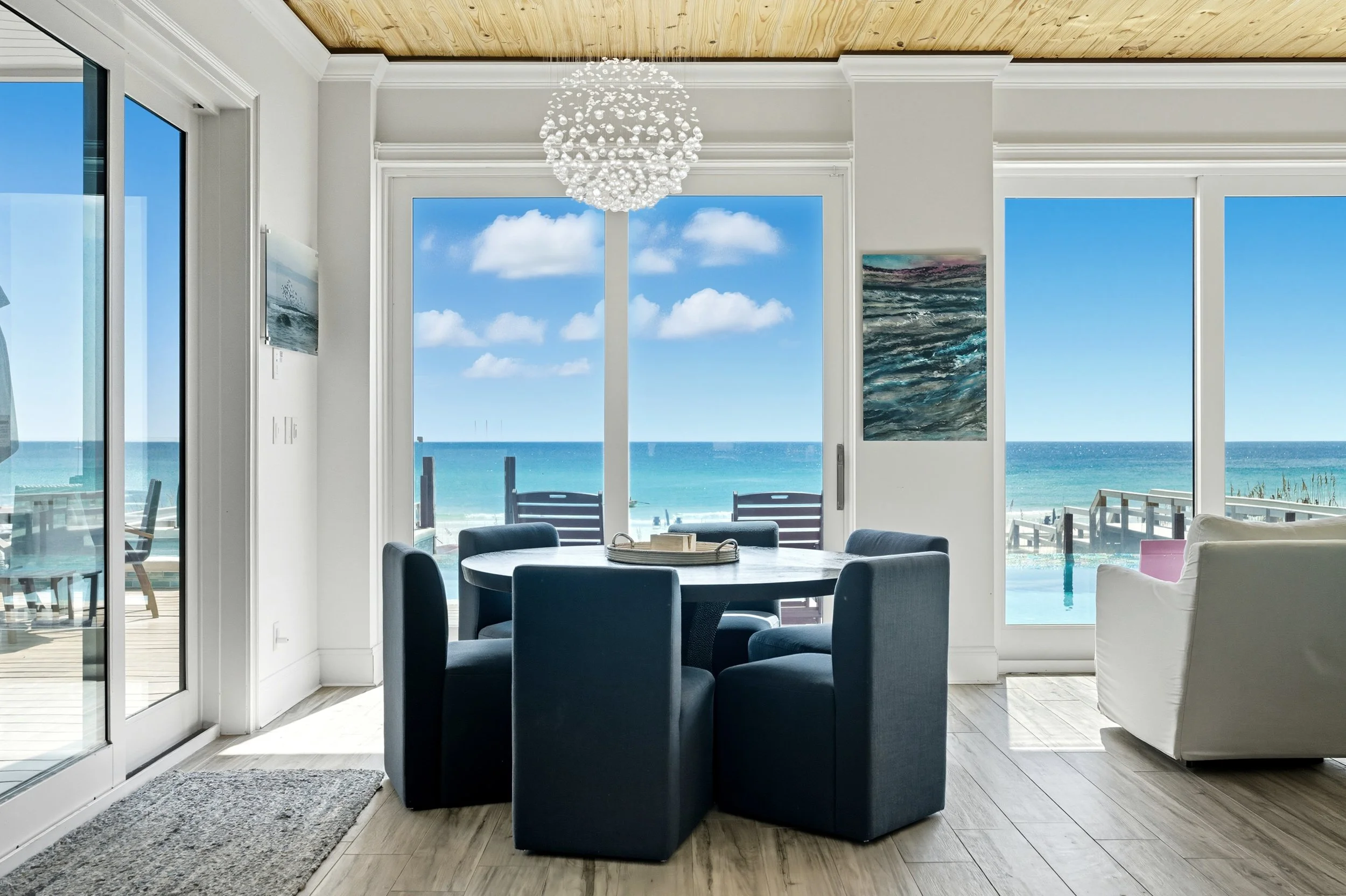 Coastal living room with large windows showing the ocean, a round dining table surrounded by six dark blue chairs, a modern chandelier, and ocean-themed artwork on the walls - Real estate photography in 30A, Destin, and Panama City Beach by Matt Jozw