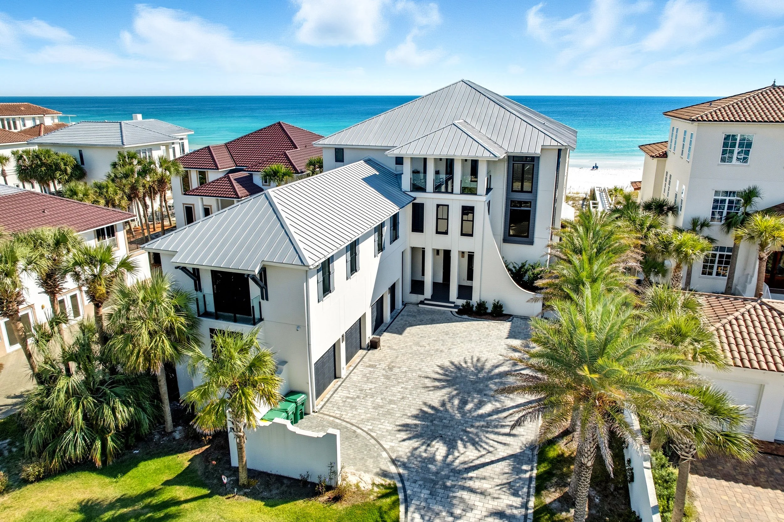 Aerial view of a modern white beach house with a metal roof, surrounded by palm trees, with neighboring houses and the ocean in the background - Real estate photography in 30A, Destin, and Panama City Beach by Matt Jozwiak
