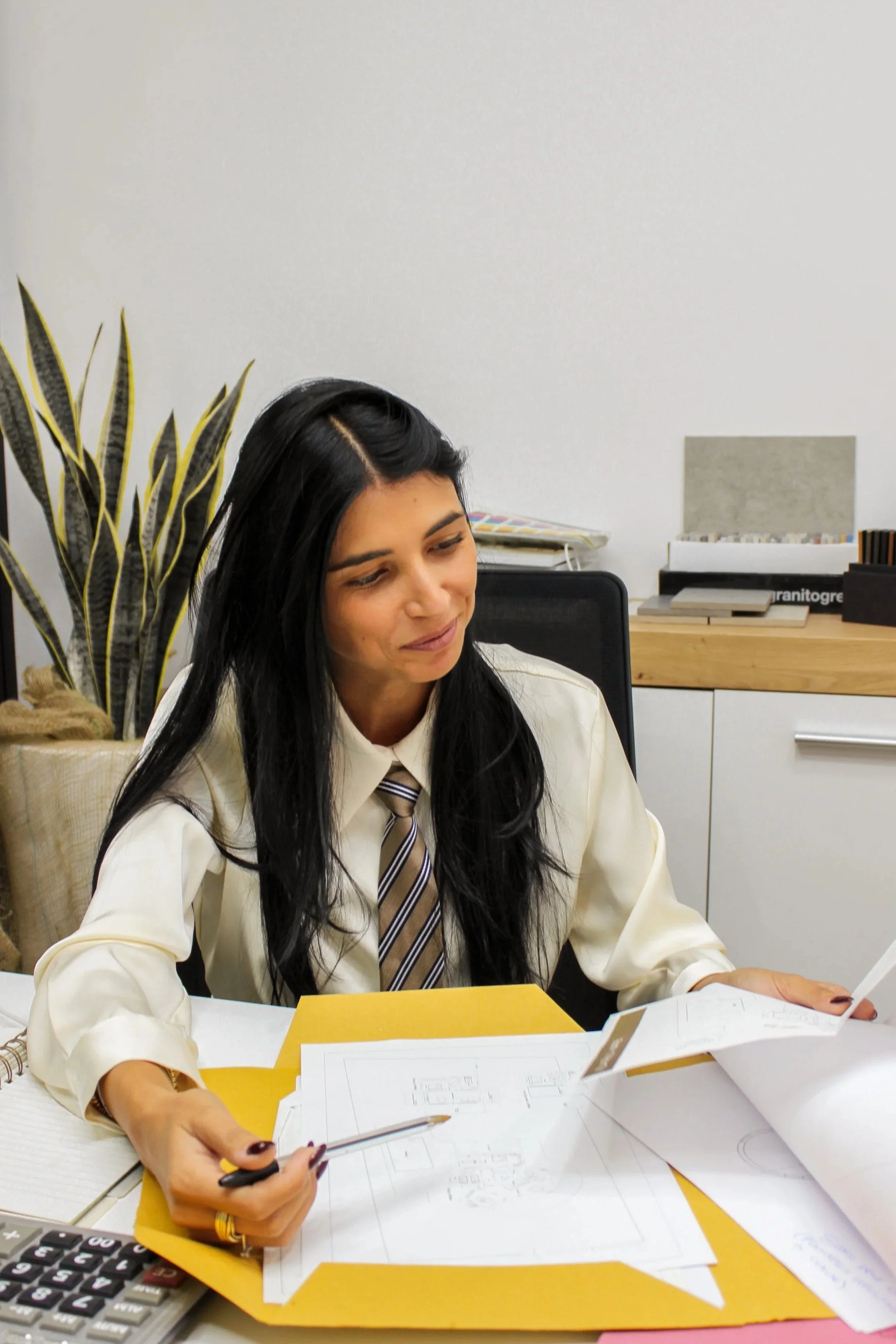 Una donna con capelli neri e abbigliamento formale seduta a una scrivania con documenti, una calcolatrice e una penna, in un ufficio con una pianta e mensole sullo sfondo.