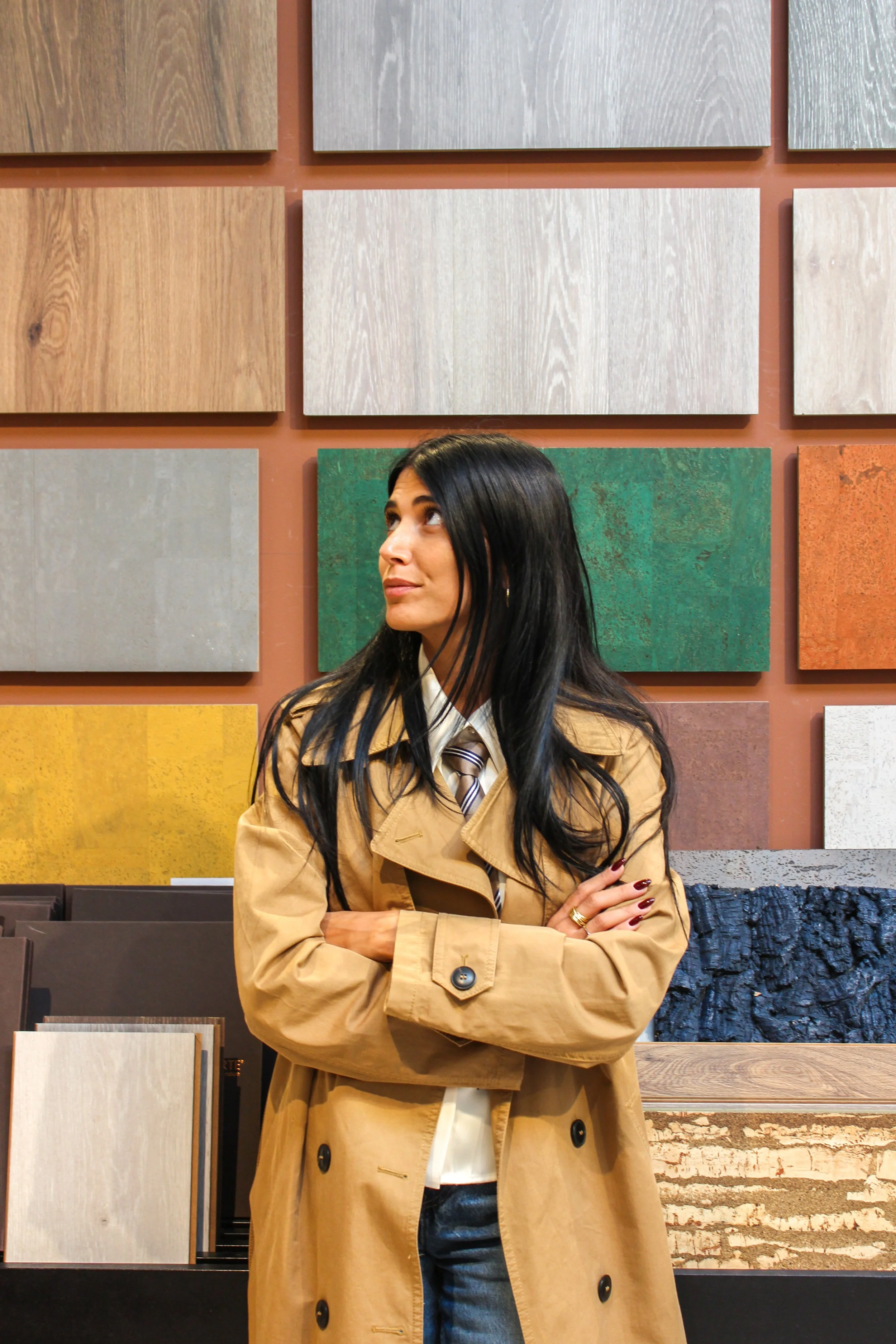 Donna con abito beige e giacca, con capelli neri, in un negozio di rivestimenti, mentre guarda i campioni di varie texture e colori alle pareti.