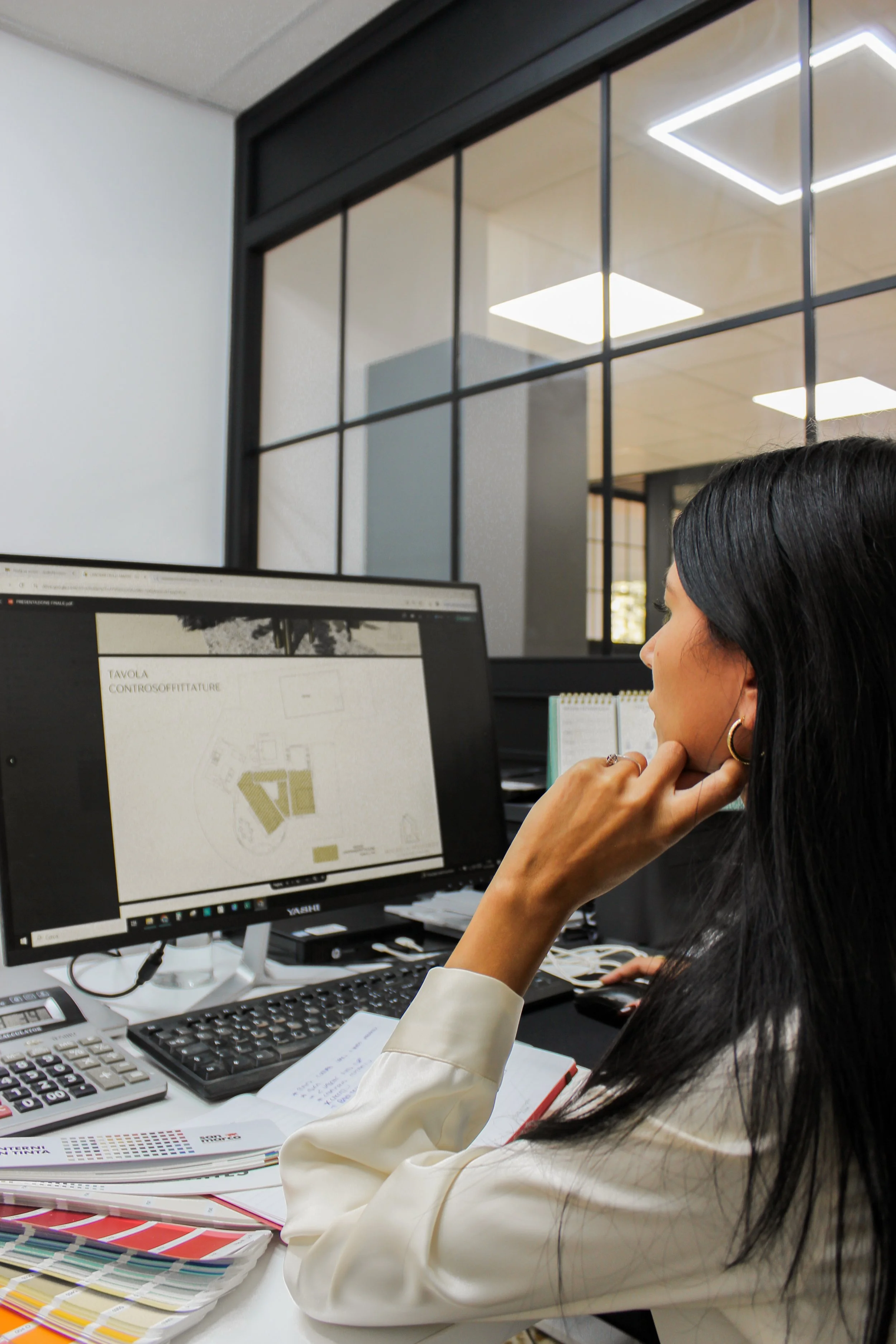 Una donna con i capelli neri, indossando orecchini d'oro e una camicia bianca, che lavora davanti a un computer in un ufficio moderno con parete vetrata.