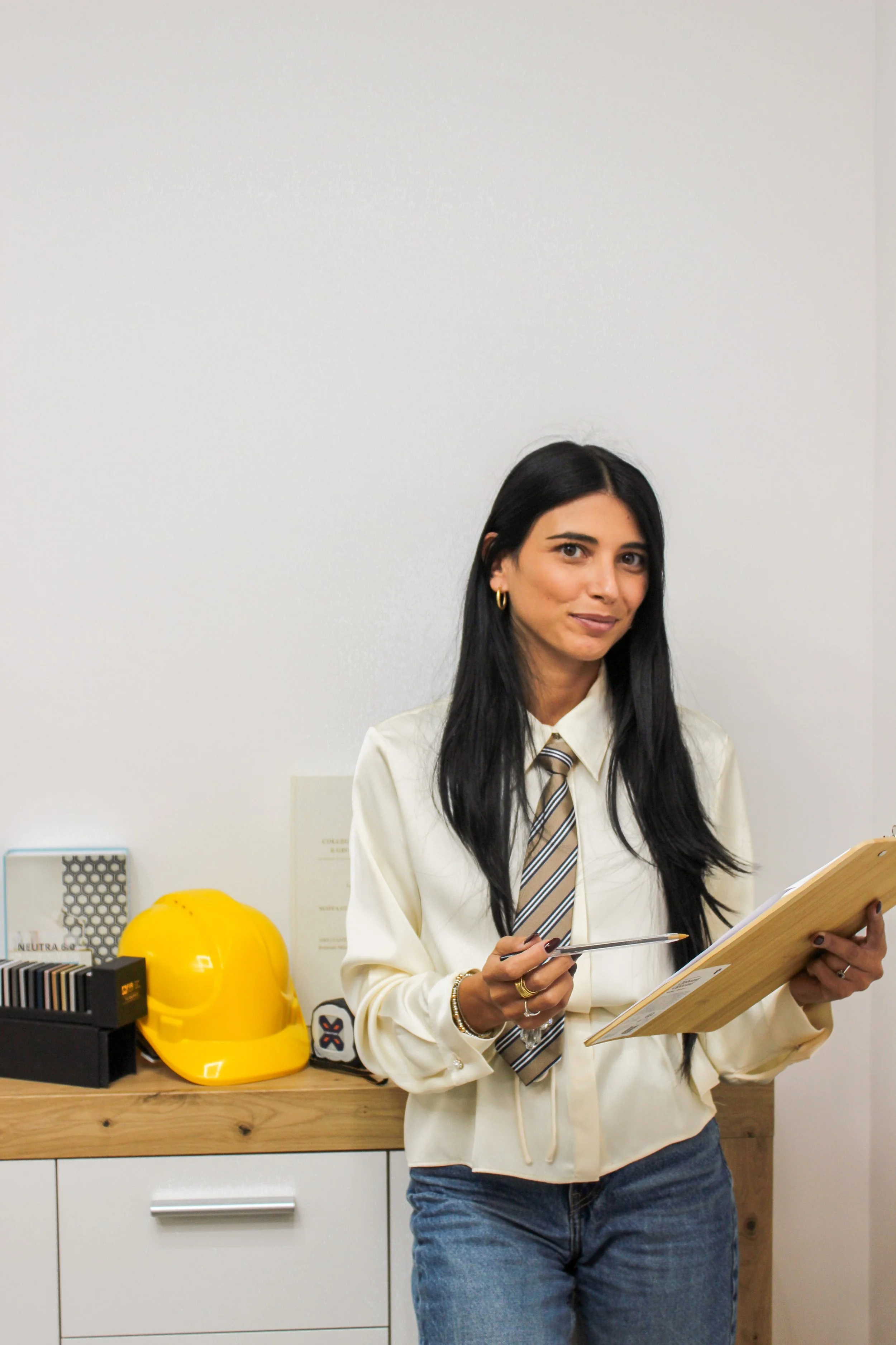una donna con i capelli neri, indossa camicia beige, cravatta a righe, e orecchini d'oro, tiene un foglio e una penna in mano, davanti a una scrivania con un casco giallo e altri oggetti, sfondo una parete bianca.