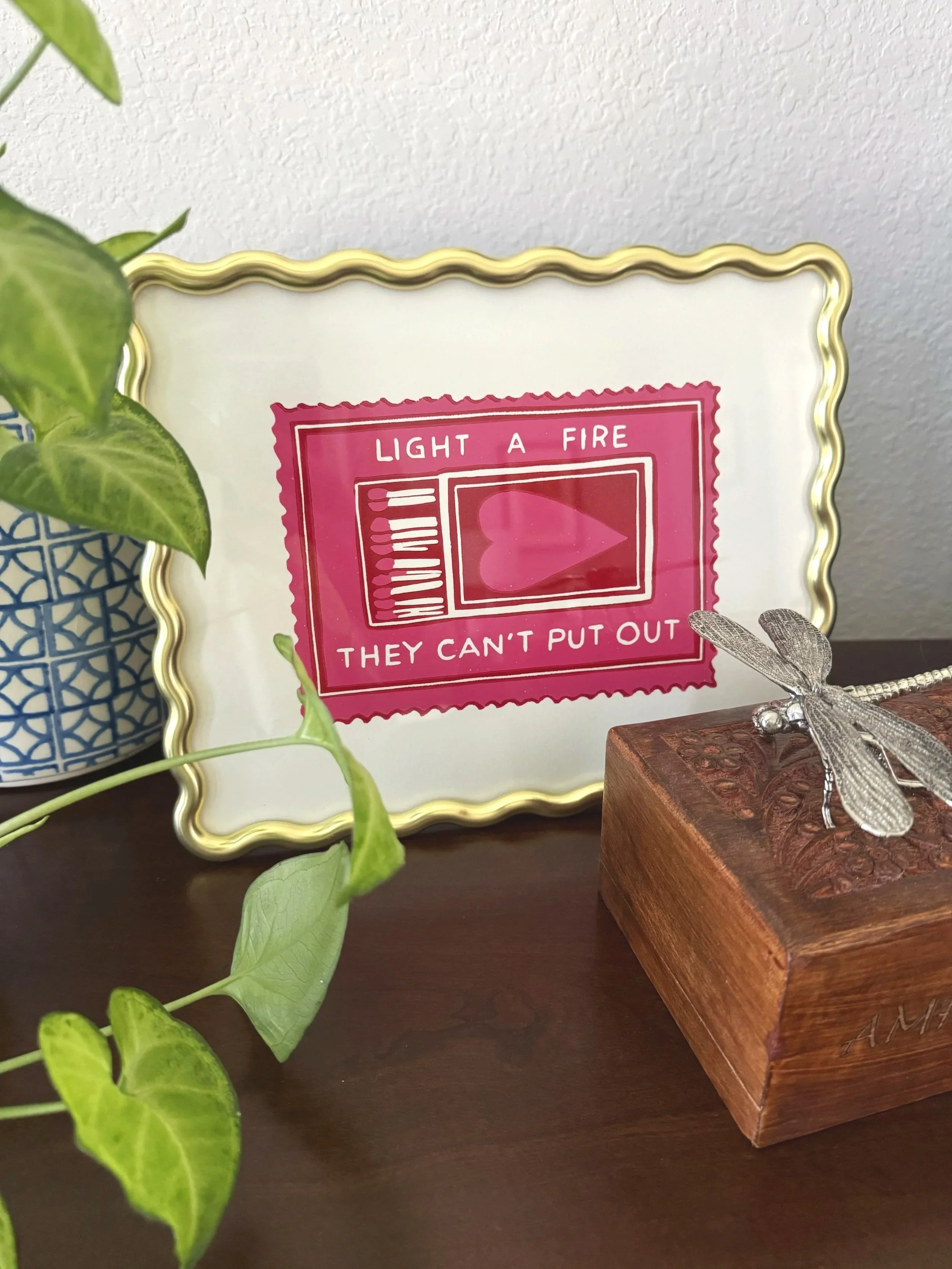 A framed poster with a pink background and white border, displaying instructions to light a fire with a humorous note that 'they can't put out.' Part of a plant with green leaves is visible on the left, and a decorative metal dragonfly figurine is perched on a small piece of wood next to a wooden box on a dark tabletop. The background is a plain white textured wall.