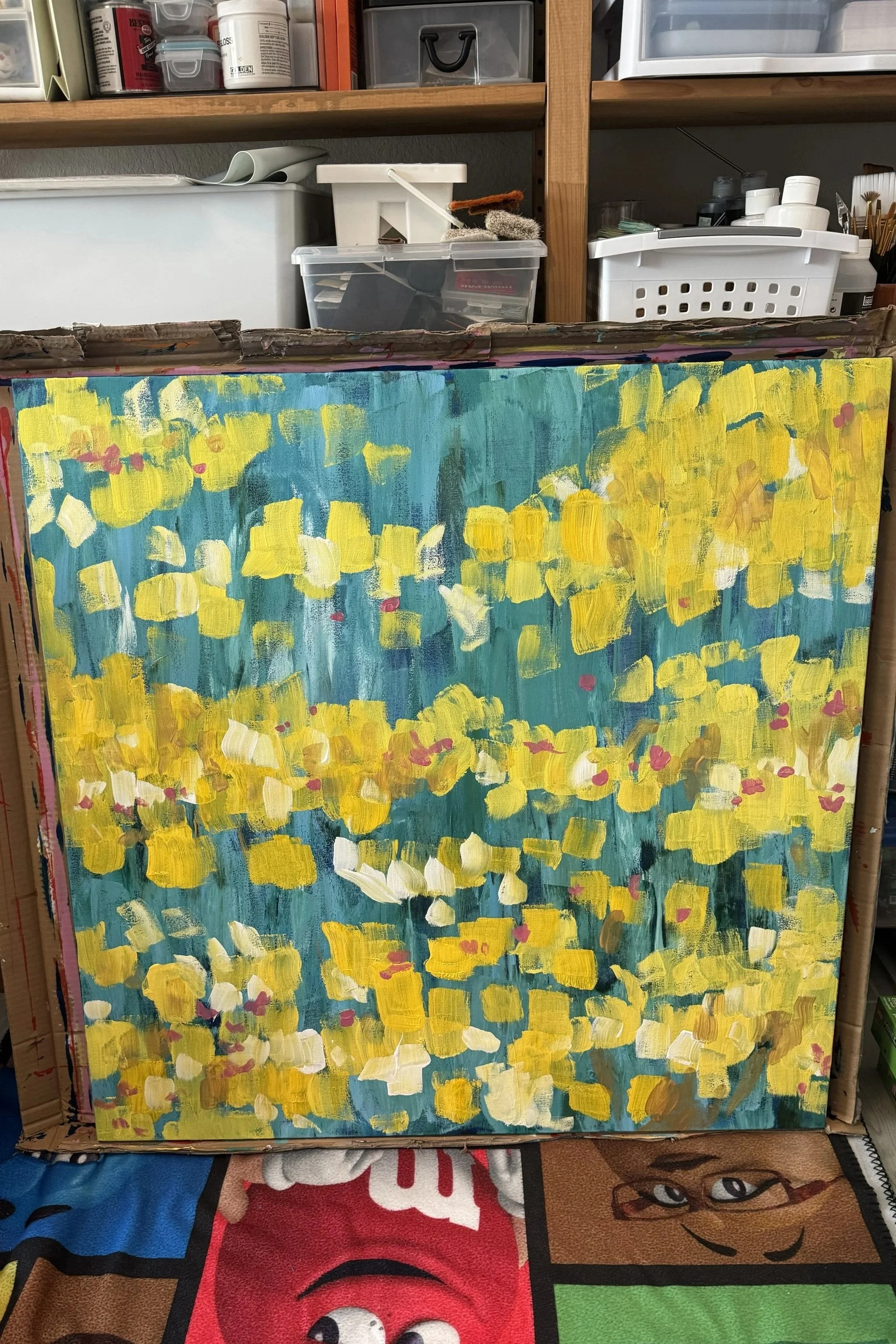 A colorful abstract painting with yellow, white, and small pink patches on a blue-green background, placed on an easel in a cluttered art studio.