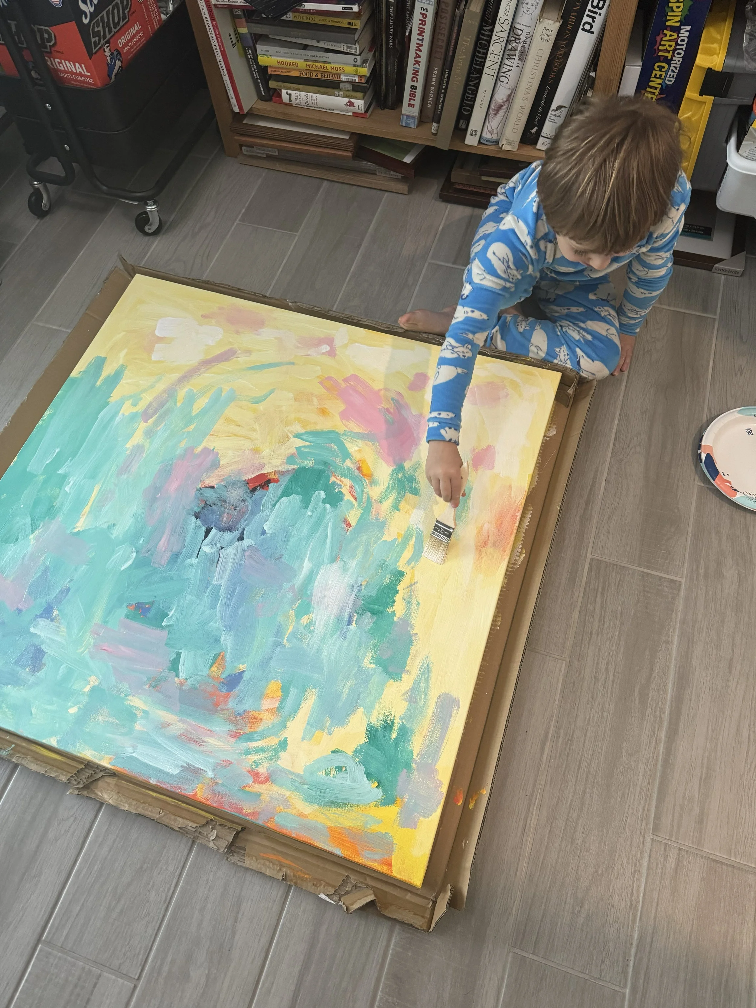 A young boy in blue pajamas with a whale pattern is crouching on a gray tiled floor, reaching out towards a large abstract painting with pastel colors on a cardboard surface, inside a room with bookshelves.