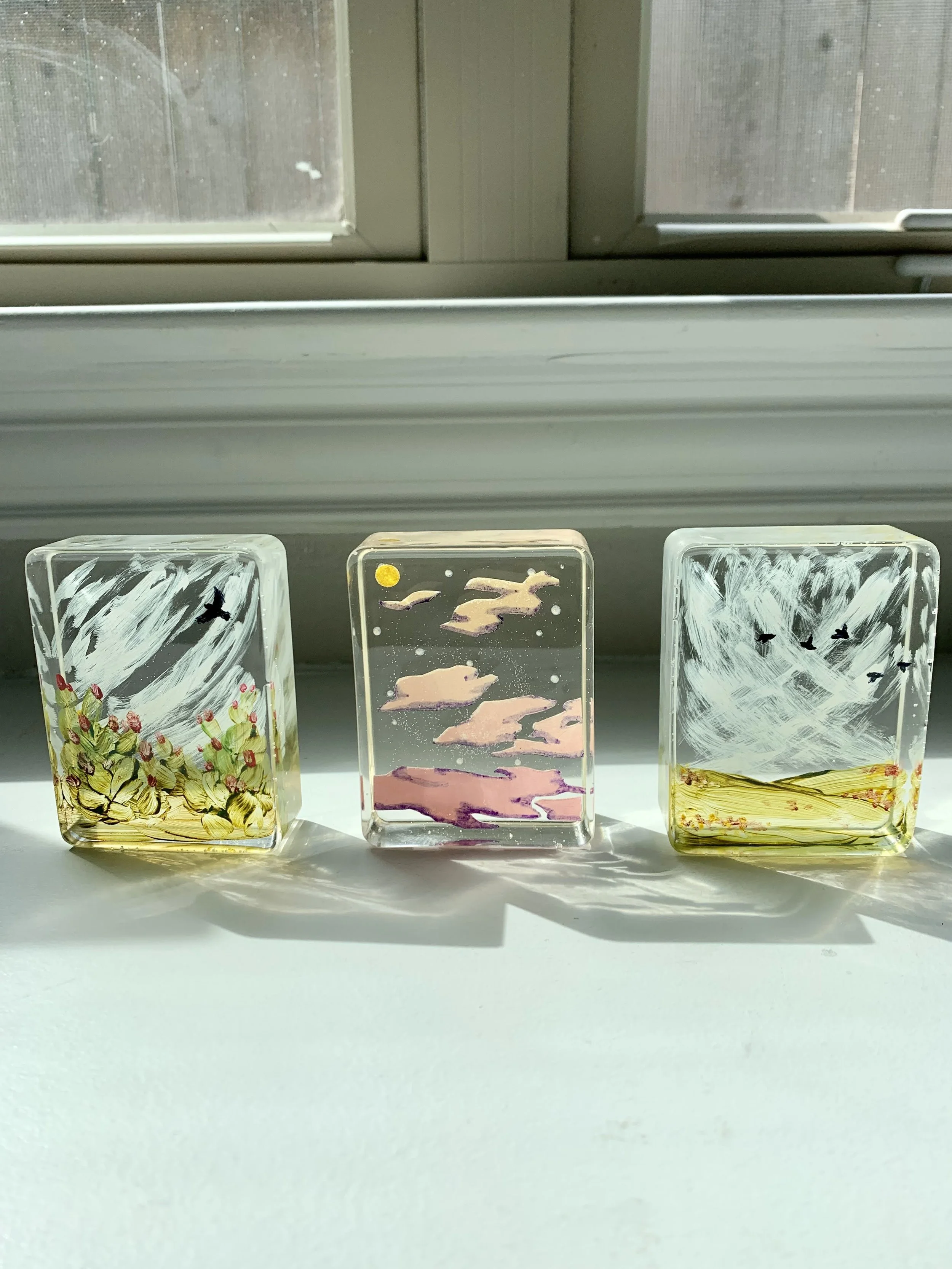 Three glass art pieces with painted designs of nature scenes, including flowers, birds, and a landscape, placed on a white surface in front of a window.
