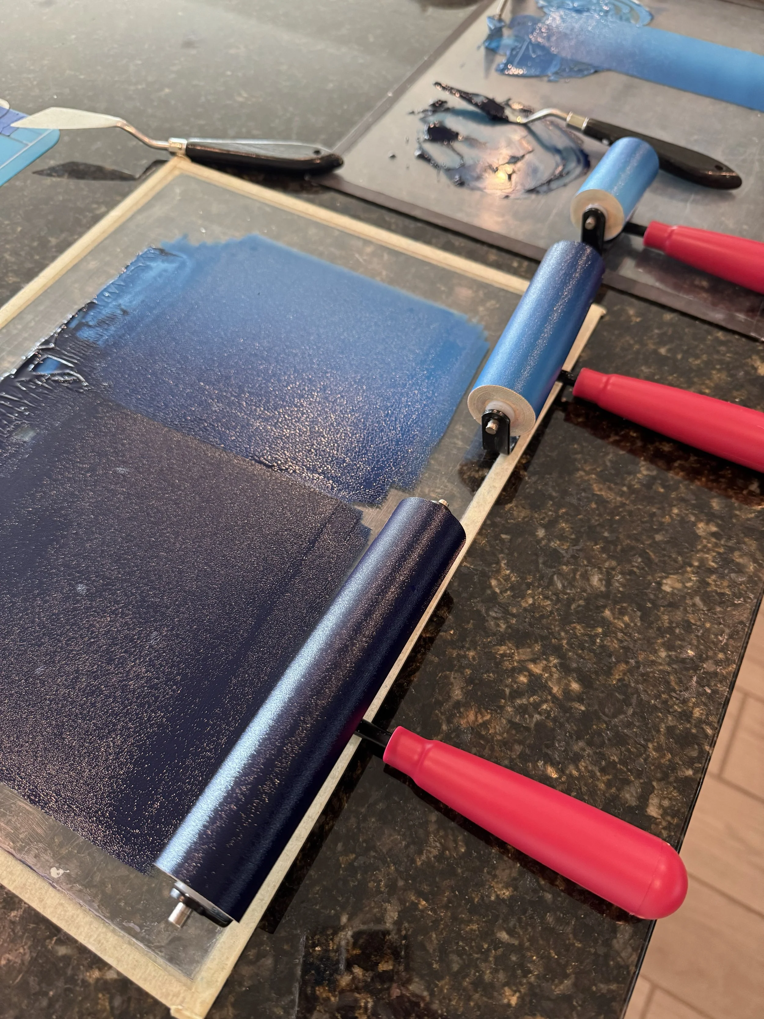 A printing or stamping device with a roller, ink, and a wooden frame, with blue ink on the roller and a sheet of blue-painted material on the surface.