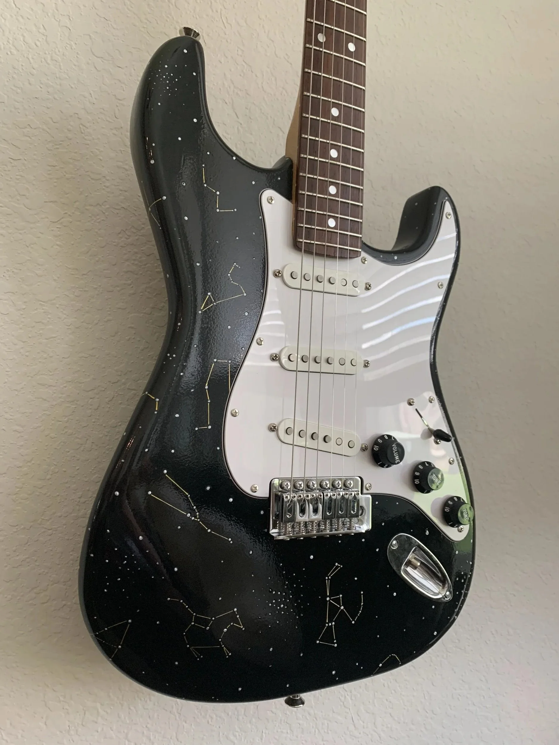 Electric guitar with a black body decorated with white constellation patterns and a white pickguard, hanging on a beige wall.
