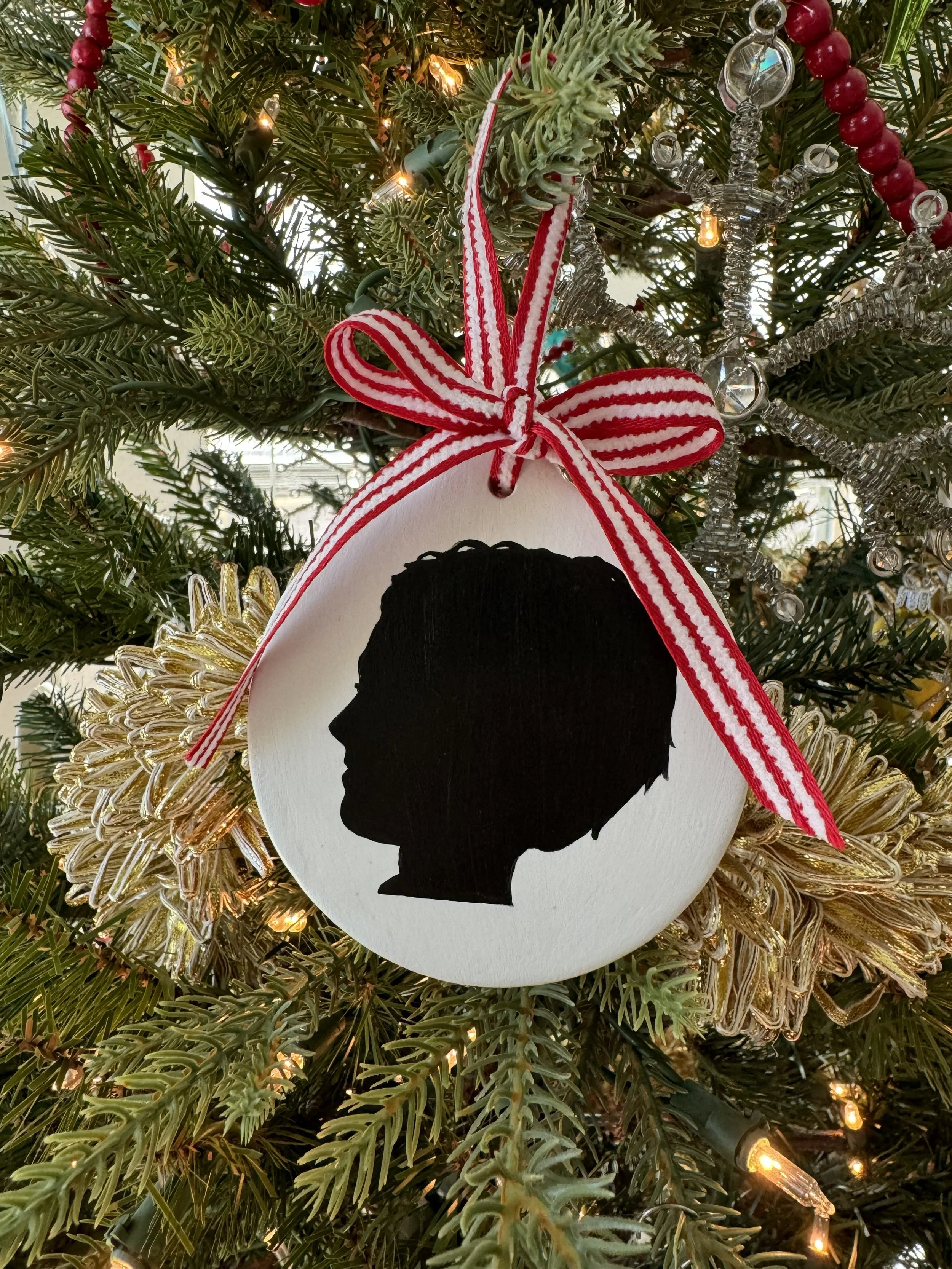 A Christmas tree ornament featuring a silhouette of a child's profile in black, hanging from a red and white striped ribbon on a decorated Christmas tree.