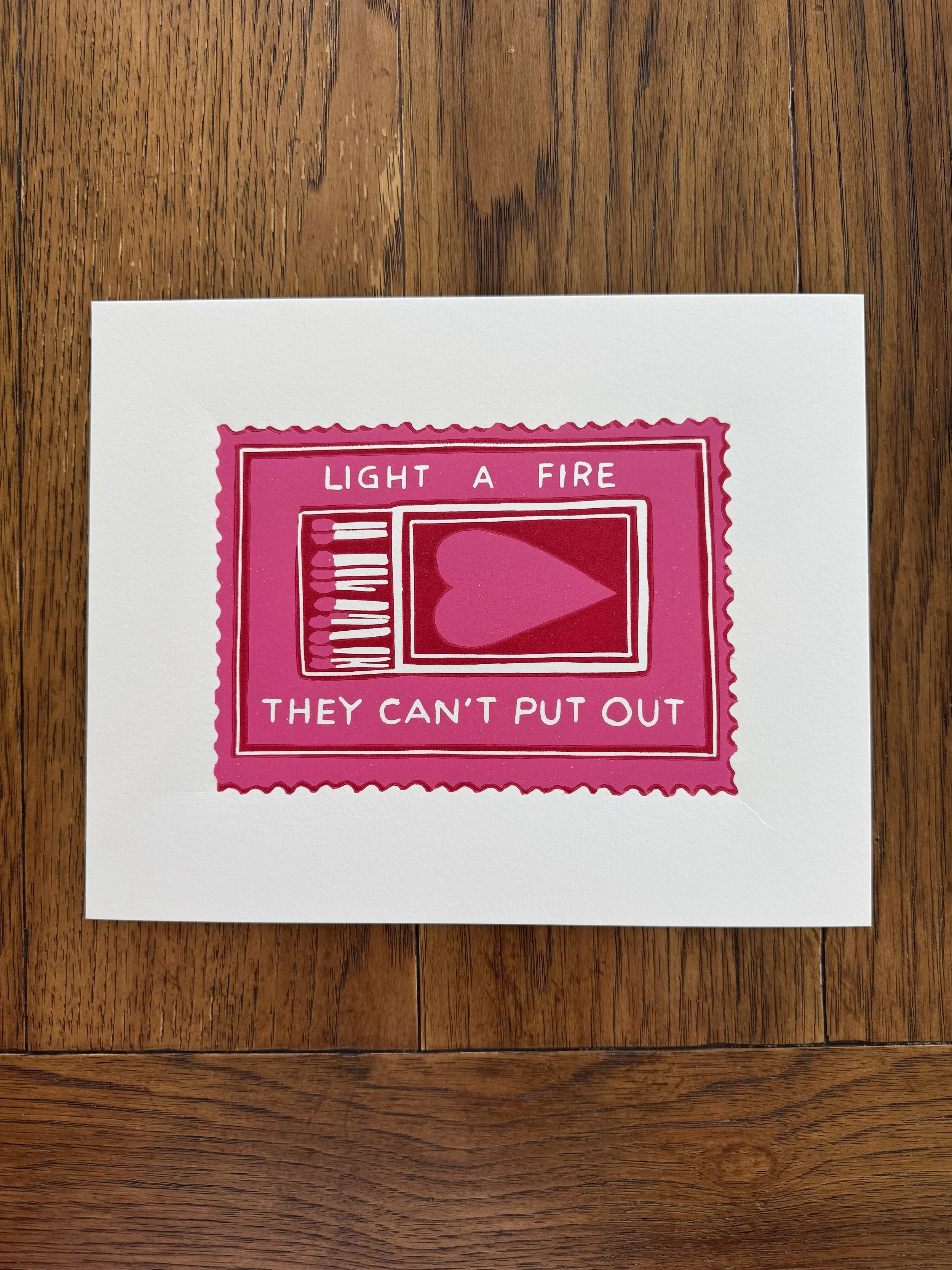 A pink postage stamp with white border and text that reads "Light a fire they can't put out," with a heart graphic in the center, on a white card on a wooden surface.