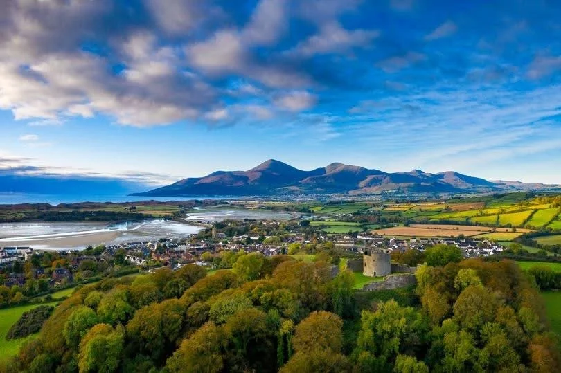 A scenic landscape with rolling green hills, a historic castle, a town, and a mountain range under a partly cloudy sky.