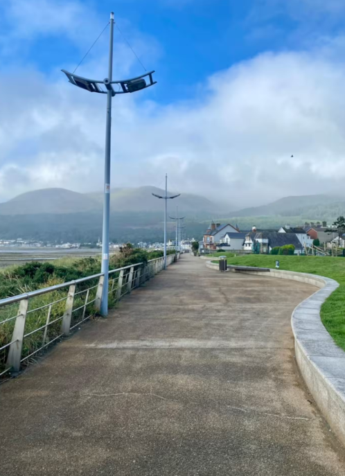 A paved walkway along a coast with modern streetlights, residential houses in the background, and hills under a partly cloudy sky.