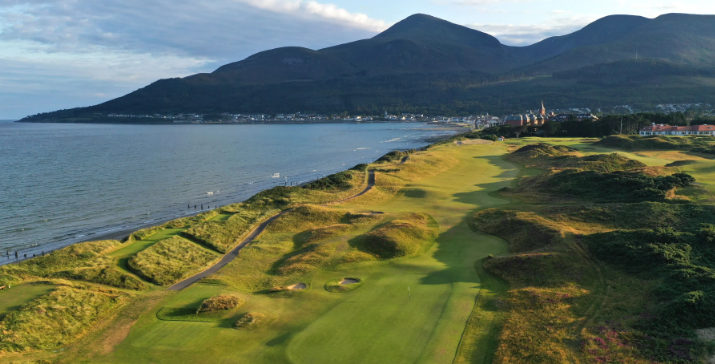 A scenic view of a coastal golf course with lush green fairways and sand traps, overlooking a body of water with mountains in the background during daytime.