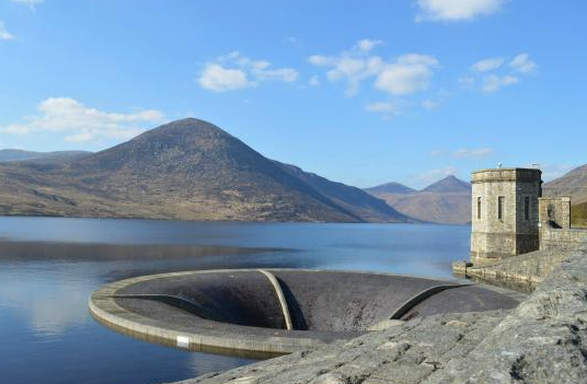 A reservoir with dam infrastructure, mountains in the background, and a partly cloudy sky.