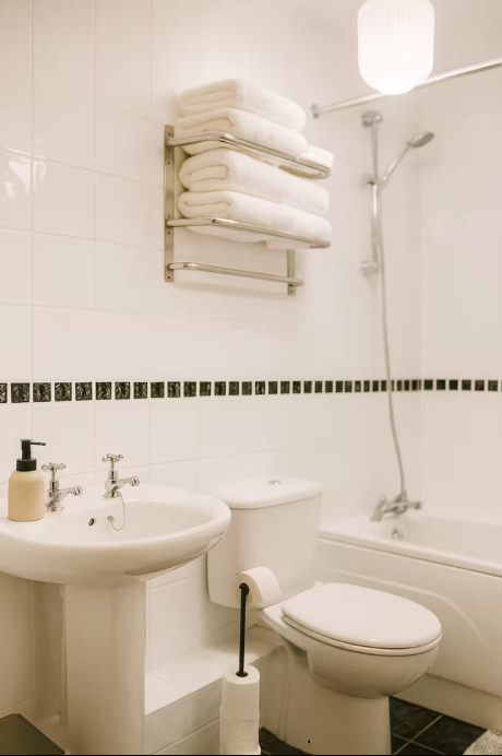 A clean white bathroom with a wall-mounted towel rack holding stacked folded towels, a white sink with a soap dispenser, a toilet, a bathtub with a showerhead, and a white hanging light fixture.