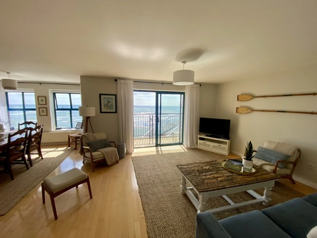 Living room with ocean view, featuring a sliding glass door to a balcony, a television, a coffee table, seating area, and dining table with chairs, decorated with nautical theme.
