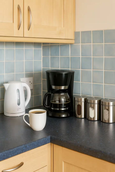 Coffee maker, electric kettle, coffee mug, and three metal canisters on a kitchen countertop with light wood cabinets and blue tiled backsplash.