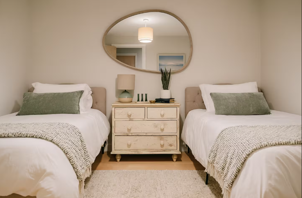 Bedroom with two twin beds, green pillows, white bedding, and a distressed white nightstand with a lamp, a plant, and decorative items, reflected in an oval mirror.