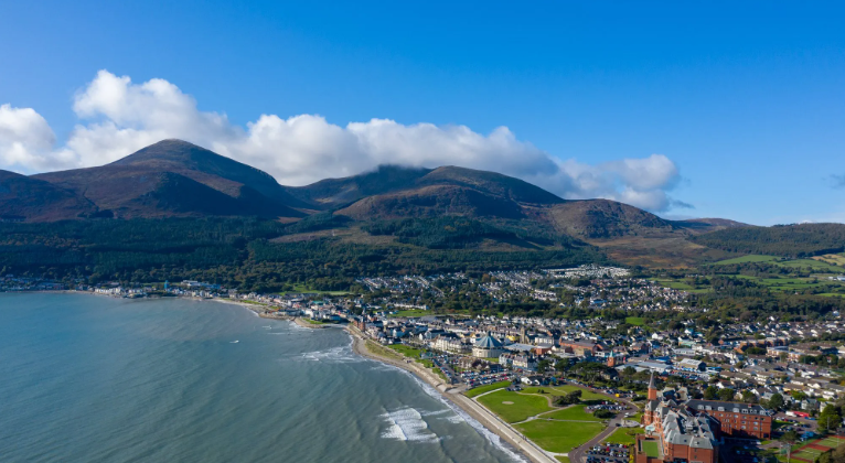 Aerial view of a coastal town with a beach, residential and commercial buildings, and green parks, with mountains in the background under a partly cloudy sky.