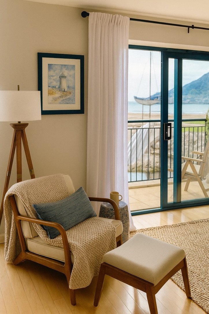 Interior of a living room with a beige armchair, a matching ottoman, a lamp, a framed lighthouse picture, and a sliding glass door leading to a balcony with outdoor furniture and a boat view.