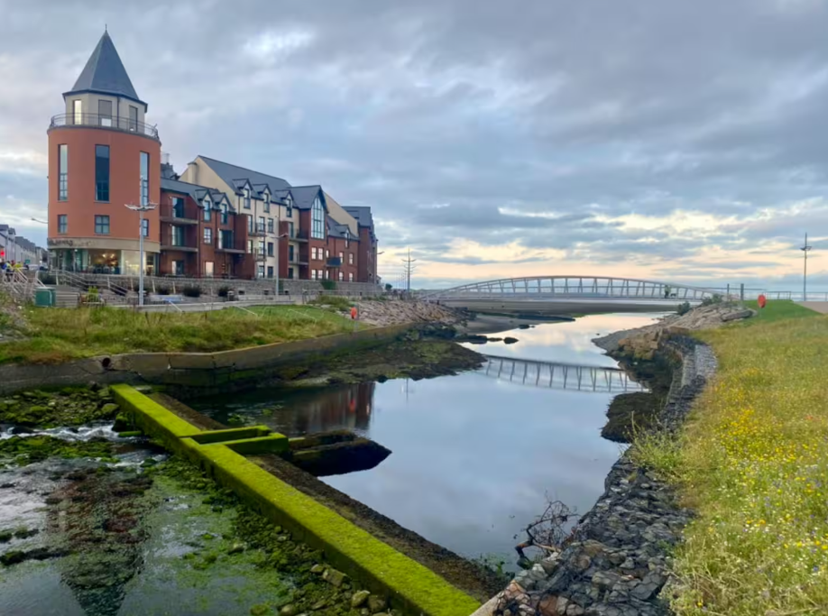 A building with a turret on the left, a small bridge in the background, and a calm body of water reflecting the cloudy sky and surroundings.