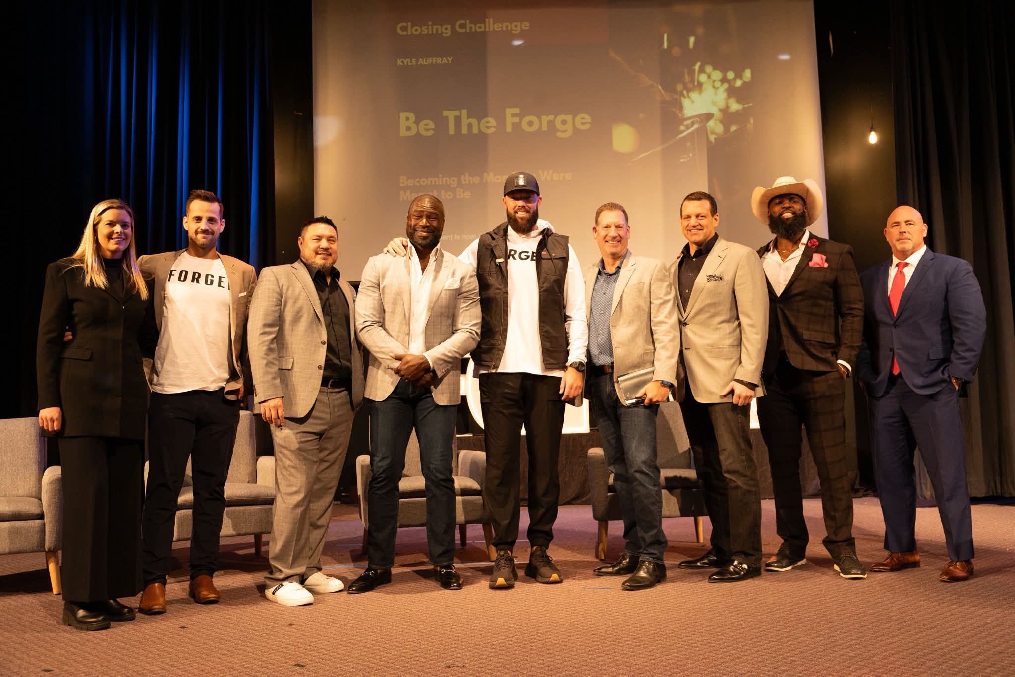 A group of ten people standing on stage in front of a screen that reads 'Closing Challenge - Be The Forge'. They are dressed in business casual and business attire, smiling for the photo.