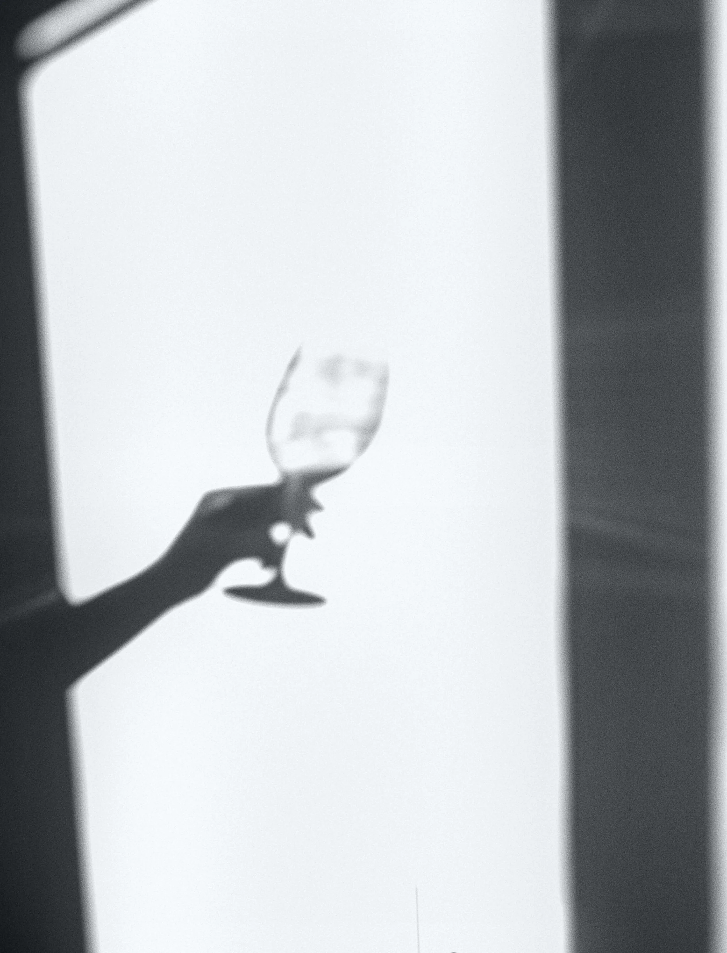 Silhouette of a hand holding a glass of wine against a bright sky with a partial shadow cast on a wall.