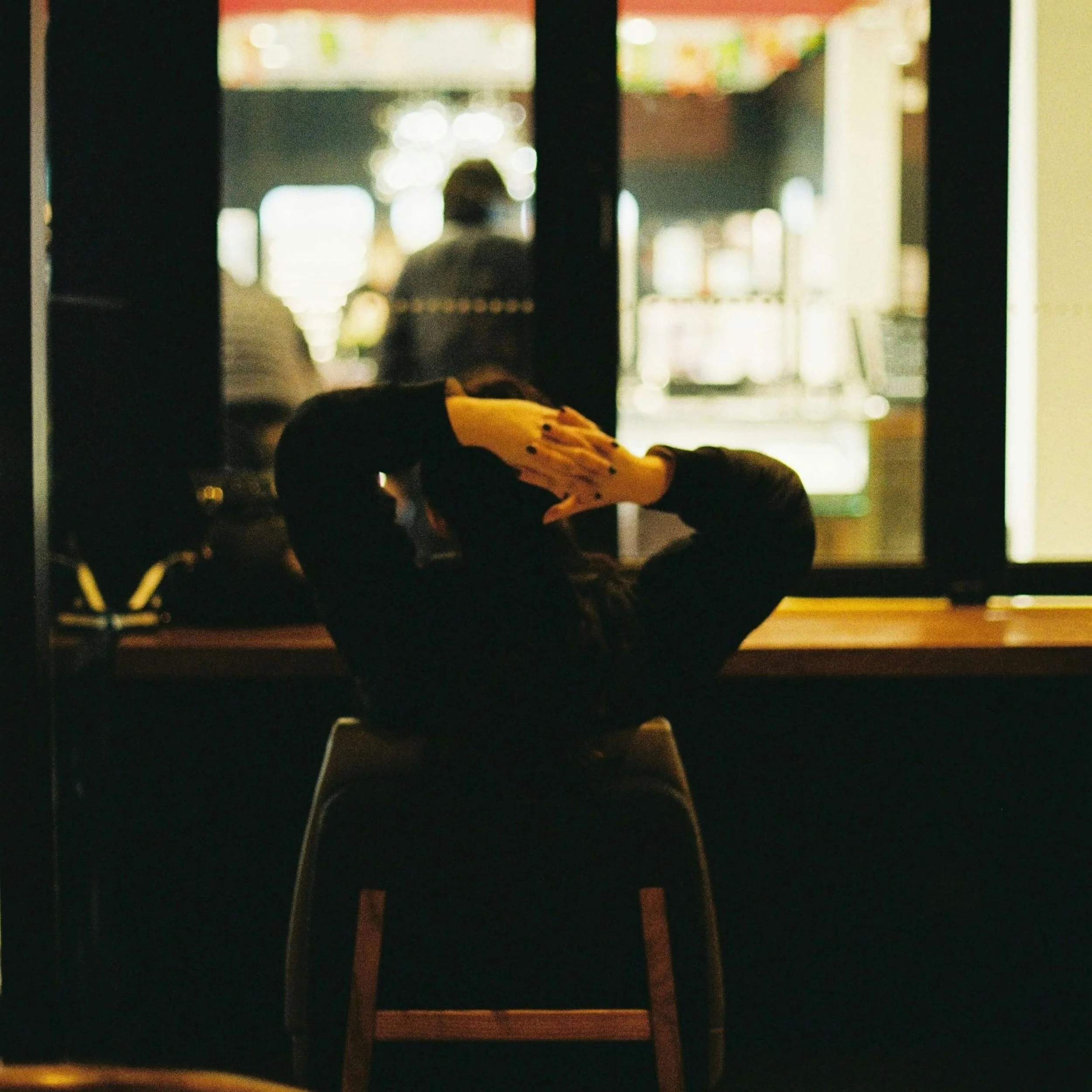 A person with dark hair sitting on a chair at a counter in a dimly lit space, resting their head and stretching their arms above their head. The background shows a brightly lit window with blurred figures and interior lights.