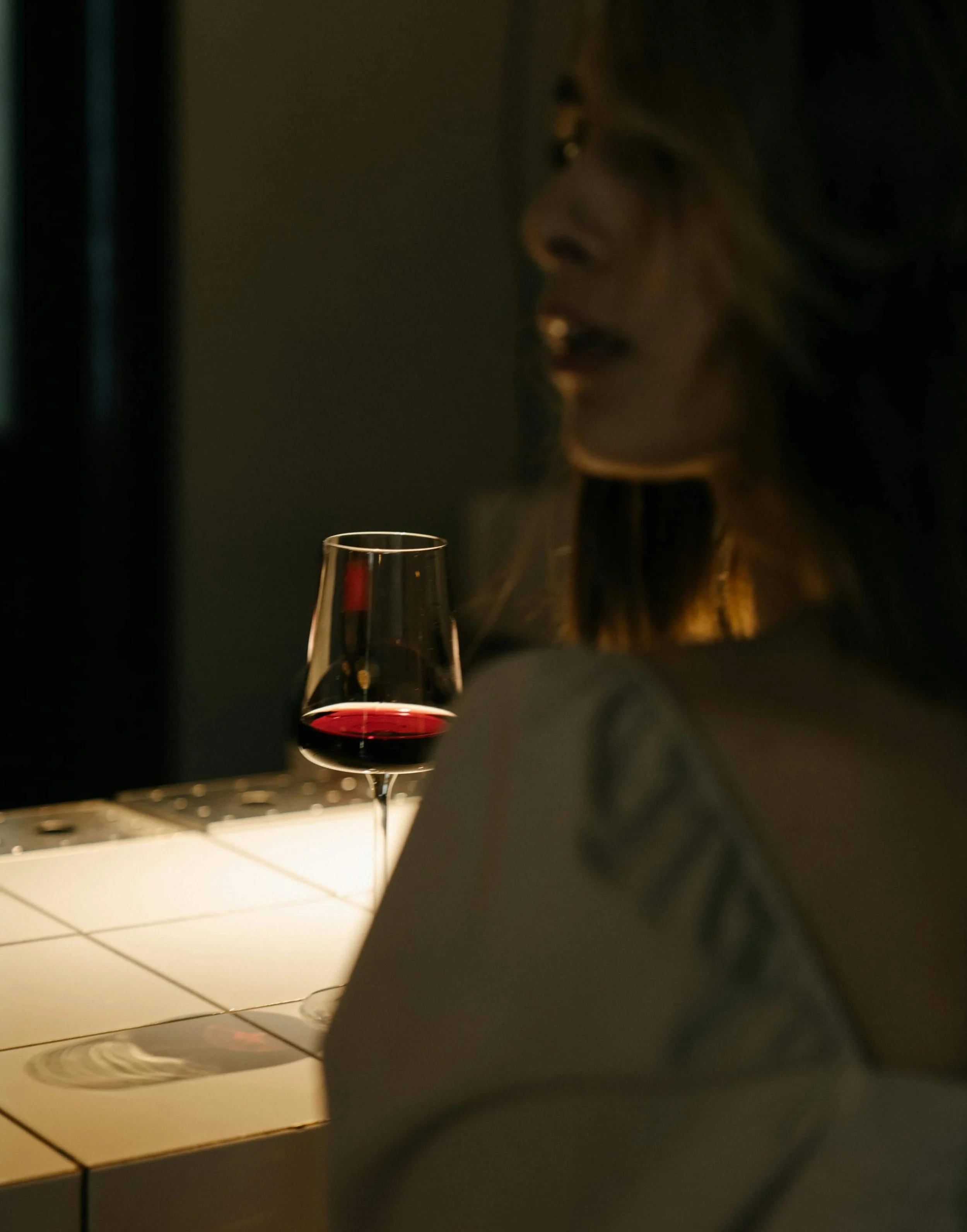 A woman with a tattoo on her shoulder sitting at a bar with a glass of red wine in front of her, dimly lit, with her face turned to the side.