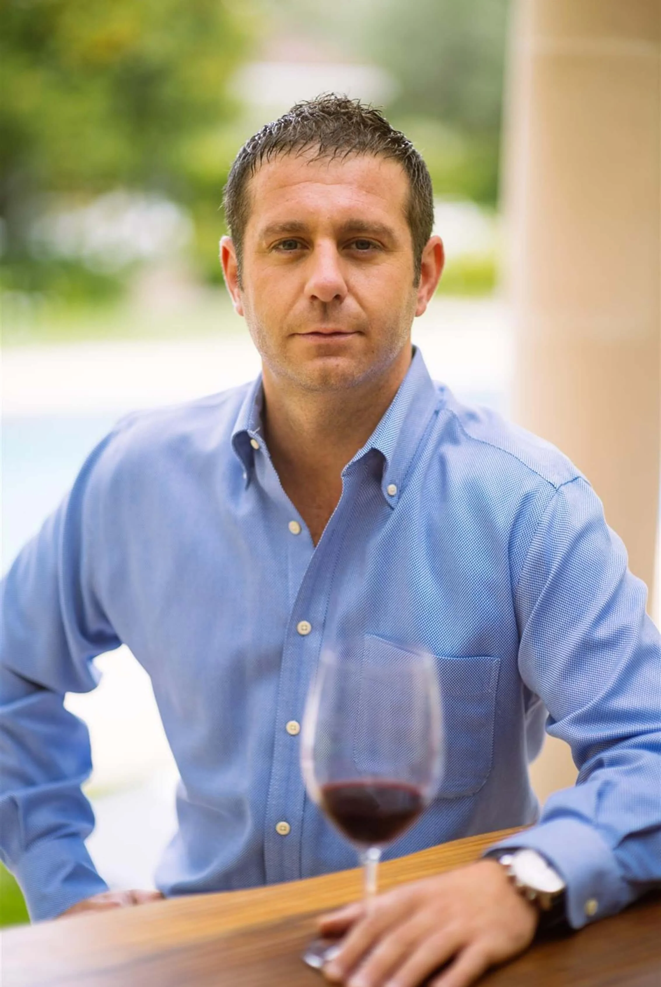 Man in blue dress shirt sitting at a table outdoors with a glass of red wine in front of him, blurred green and beige background.