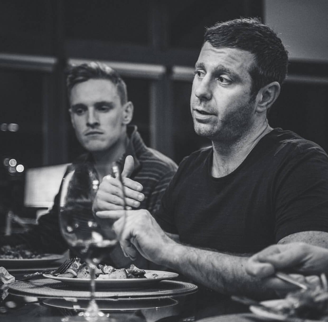 Two men at a dinner table, one in the foreground talking and the other in the background looking serious, with wine glass and plates of food.