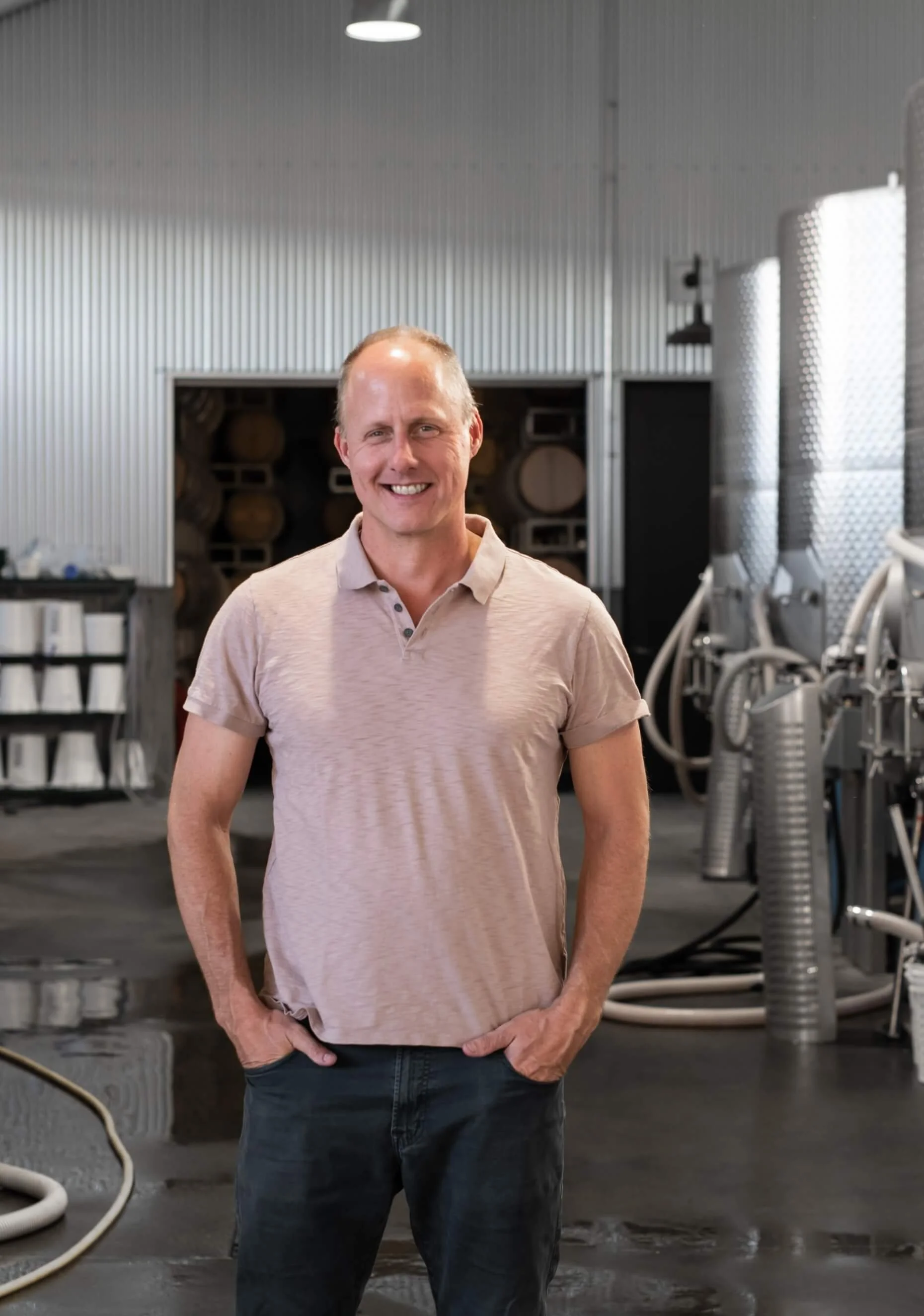 A man smiling with hands in pockets stands in a brewery or industrial setting with barrels and brewing equipment in the background.
