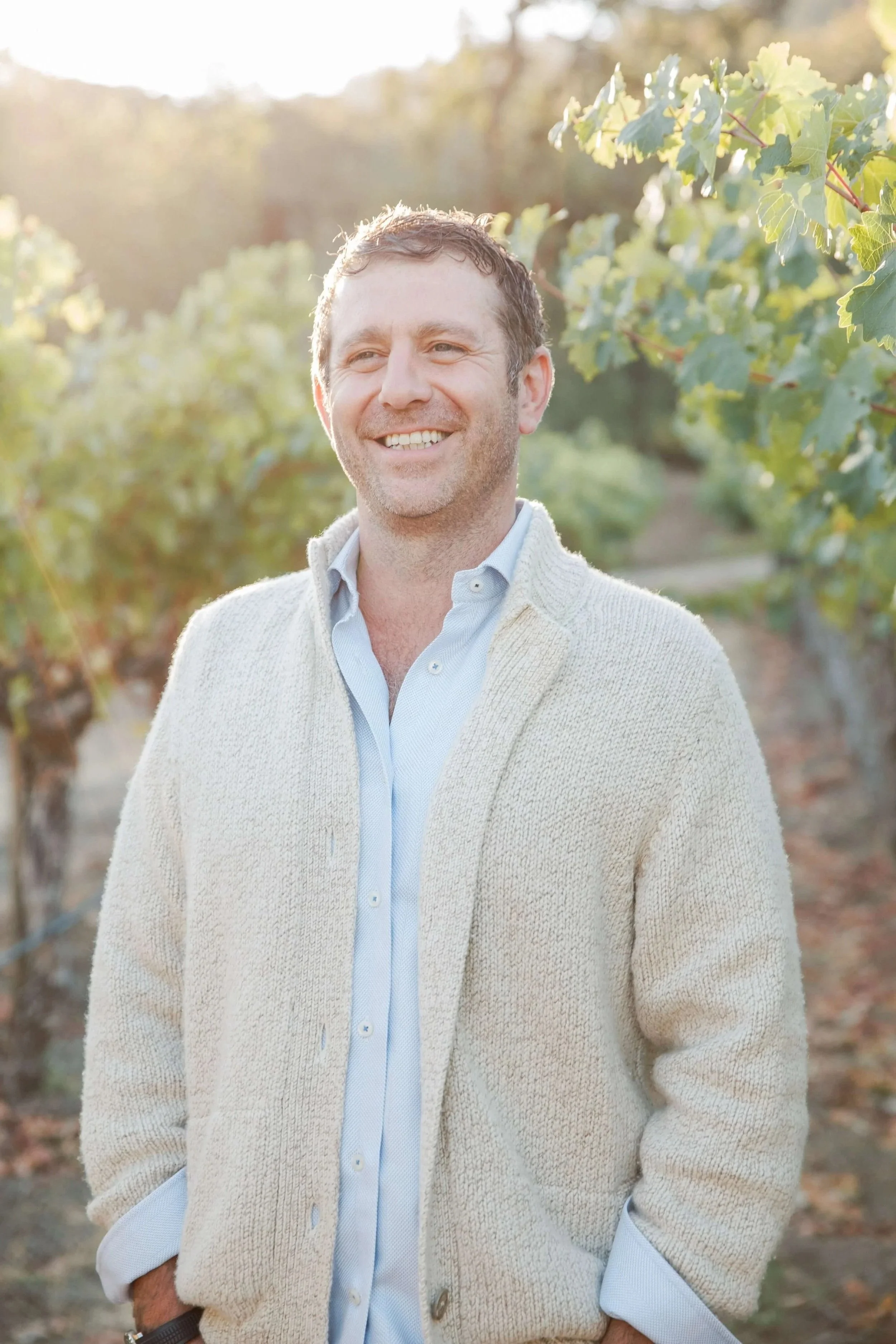 Smiling man in a sweater over a collared shirt standing in a vineyard during sunset.