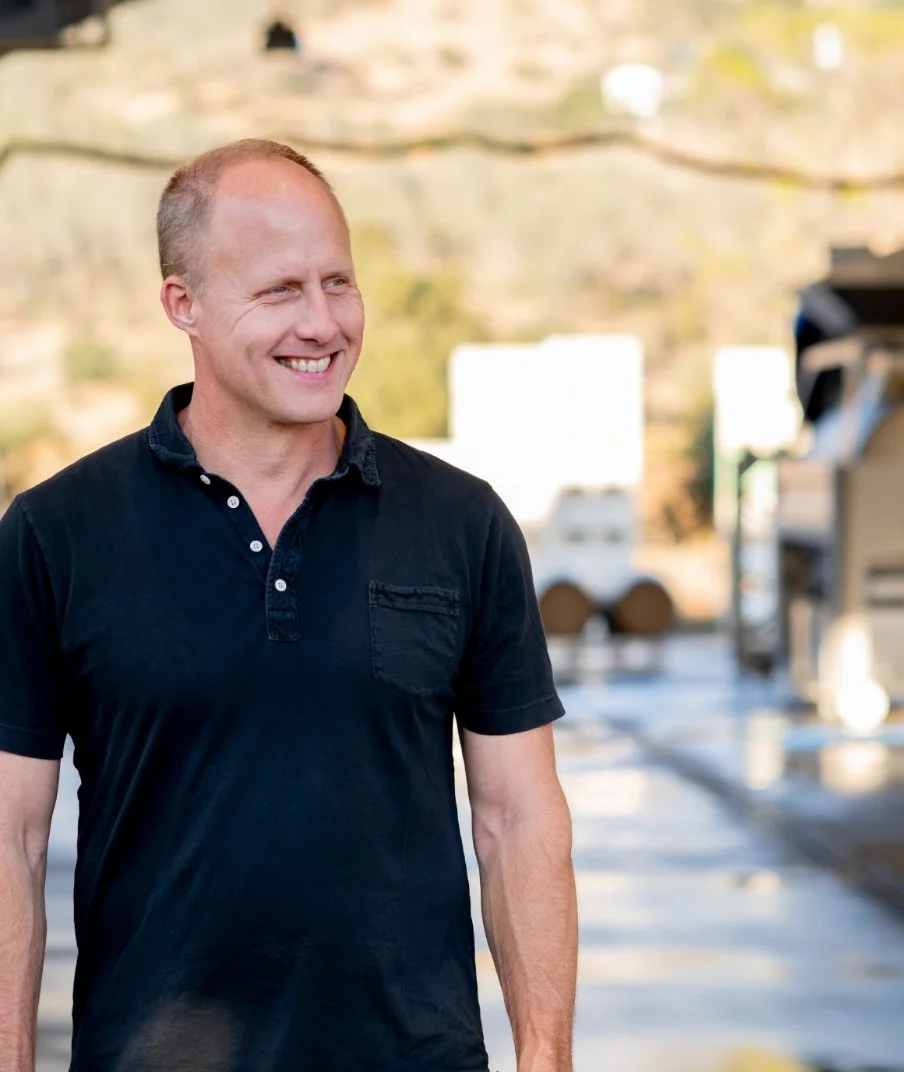 A man with a shaved head and a broad smile, wearing a dark short-sleeved polo shirt, standing outdoors with blurred vehicles and a screen in the background.