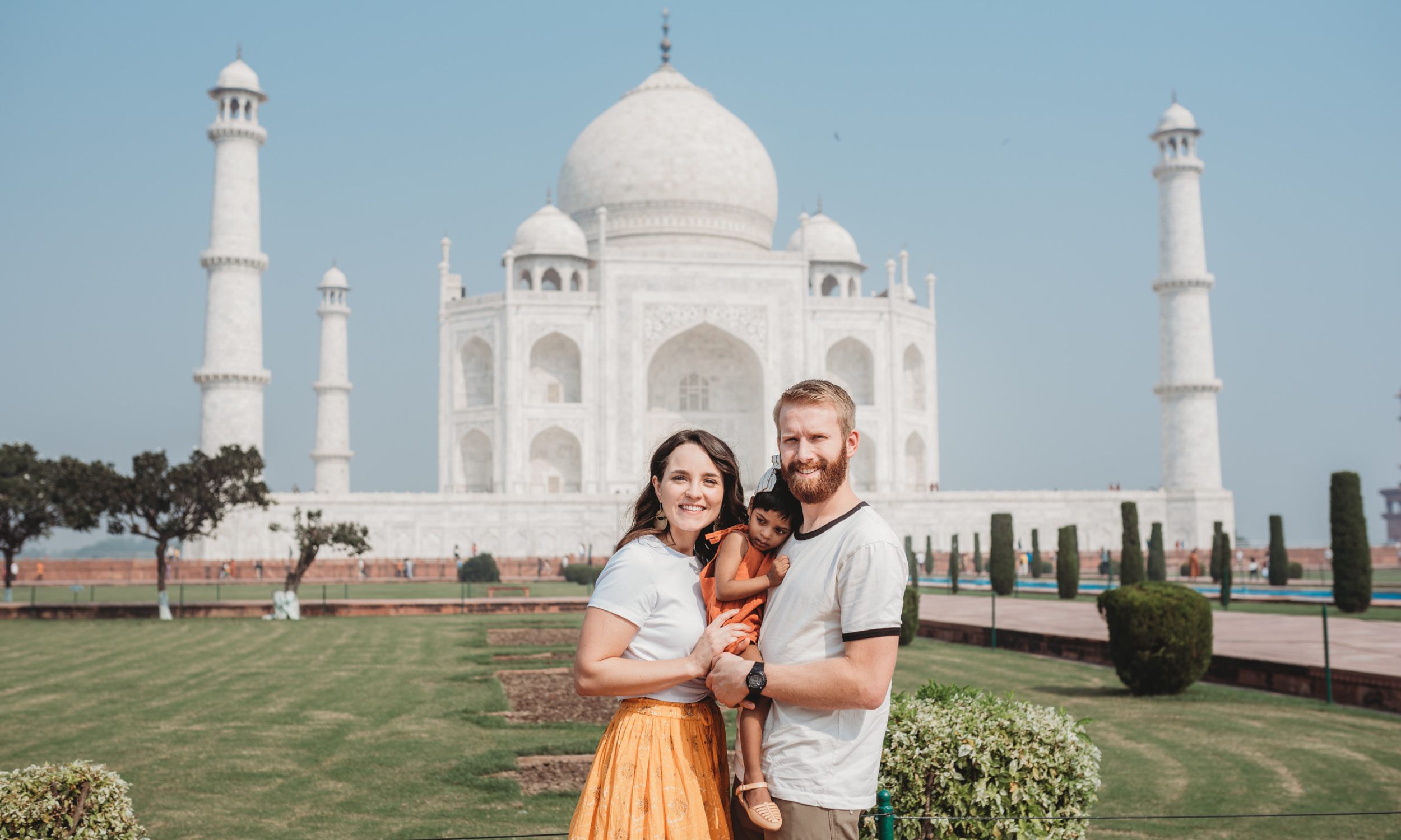 Adoptive family of three featuring India adoption travel by America World. They are standing in front of the Taj Mahal, with the mother, father, and young girl smiling at the camera.