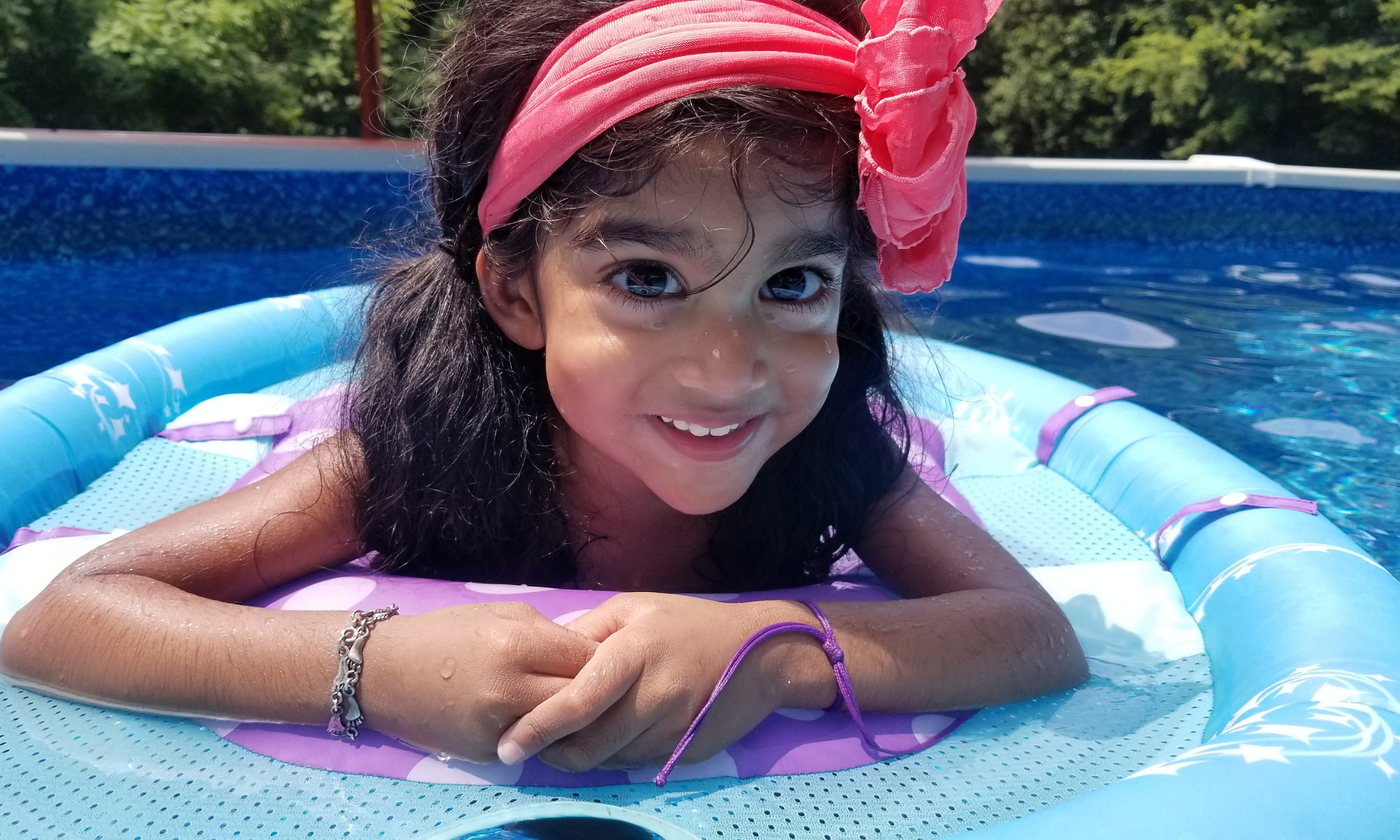 Young Indian girl with dark hair, wearing a pink headscarf, lying on a small pool float in a swimming pool, smiling at the camera.