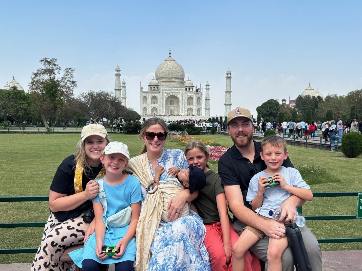 A family of six posing in front of the Taj Mahal in India to feature adoption travel with America World. The family includes three children, a man, and two women, all smiling.