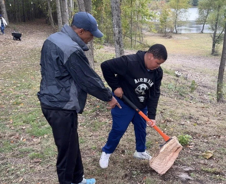 A young boy chopping wood with an axe while an older man supports him, outdoors in a wooded area near a lake.