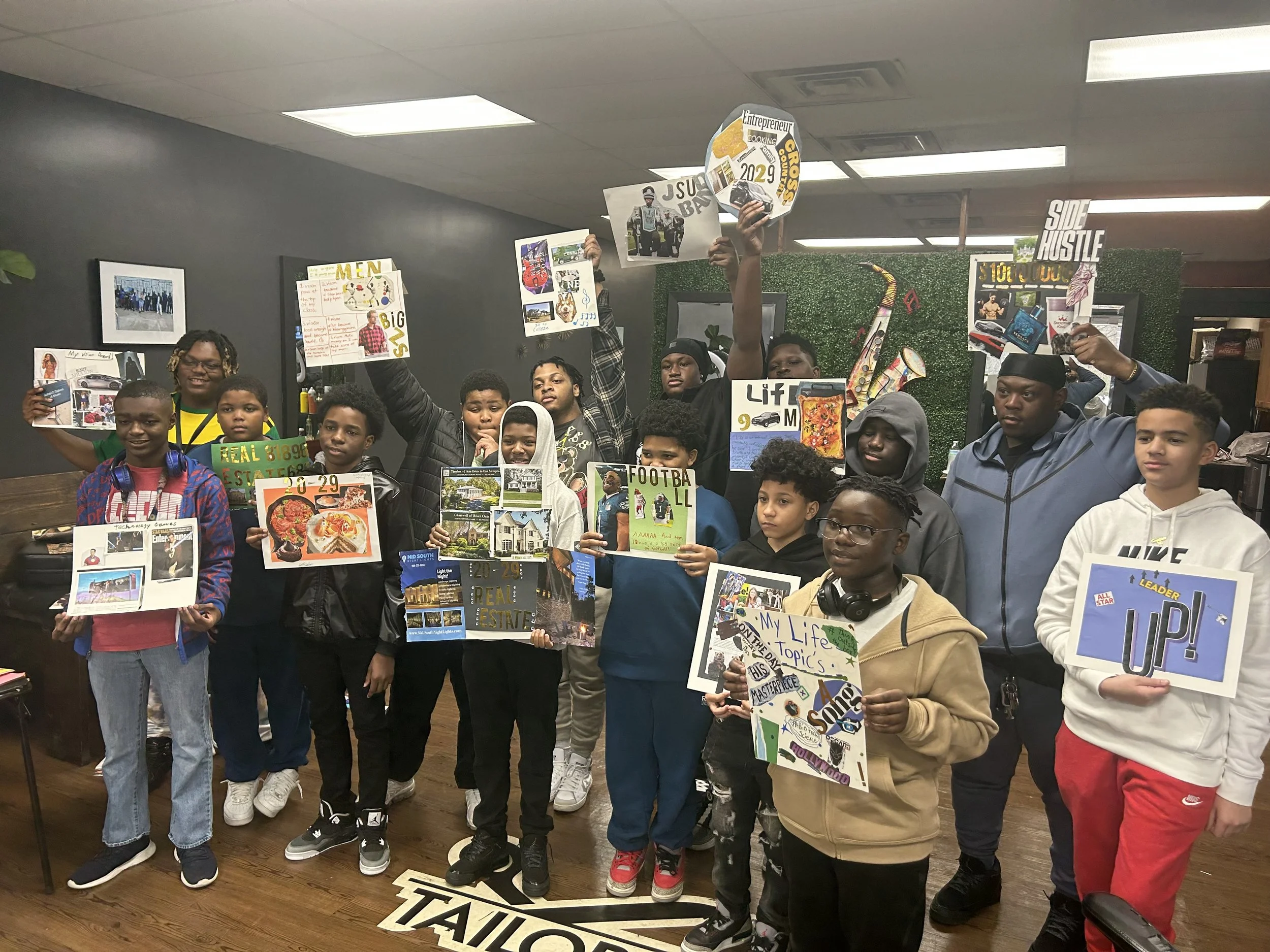 Group of children and adults inside a room, holding posters and signs with various messages, such as 'My Life Topics,' 'Lift,' and 'Up!' in a classroom or community center setting.