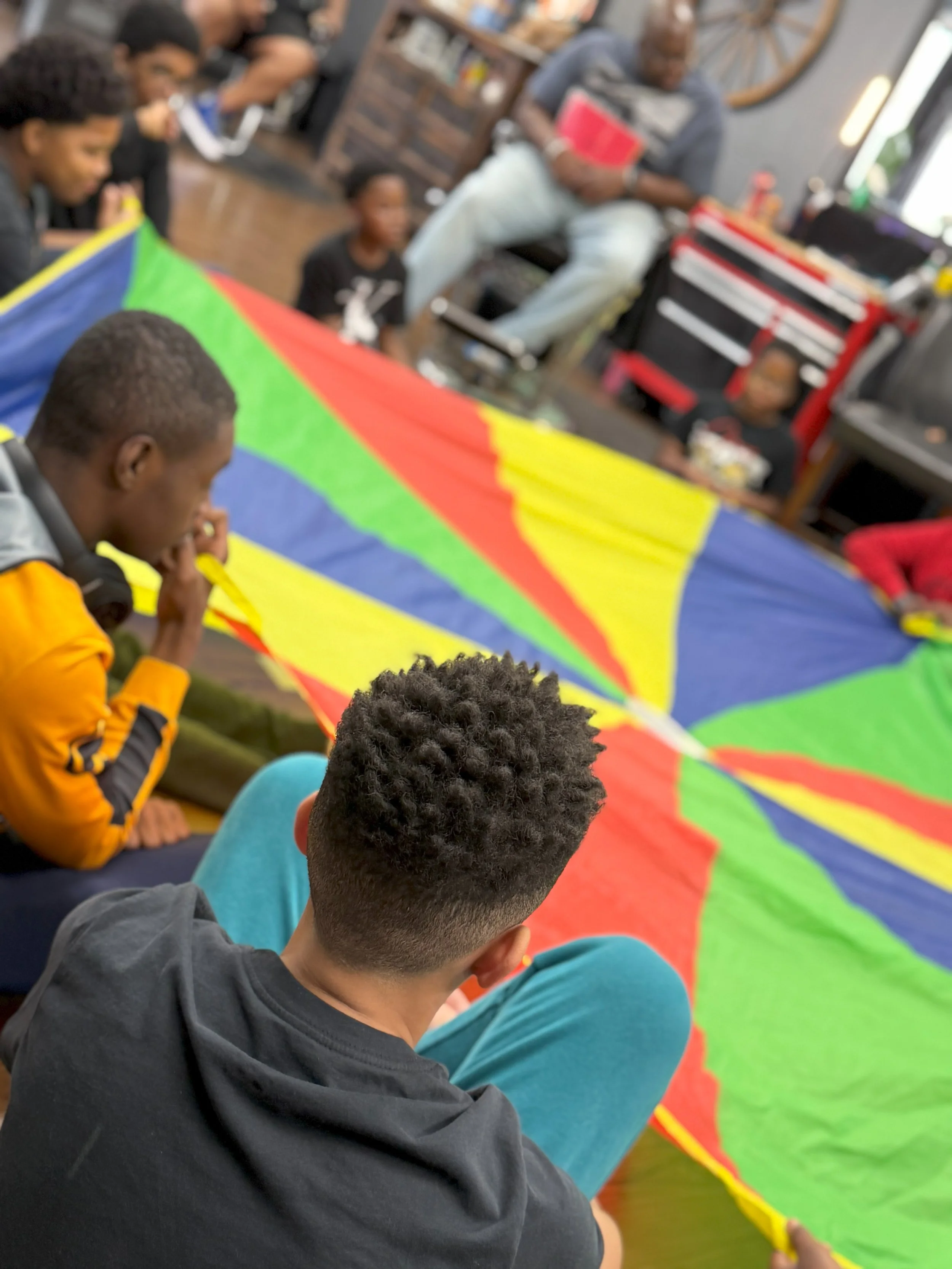 Group of children and adults sitting around a colorful parachute inside a room with wooden floors and shelves.