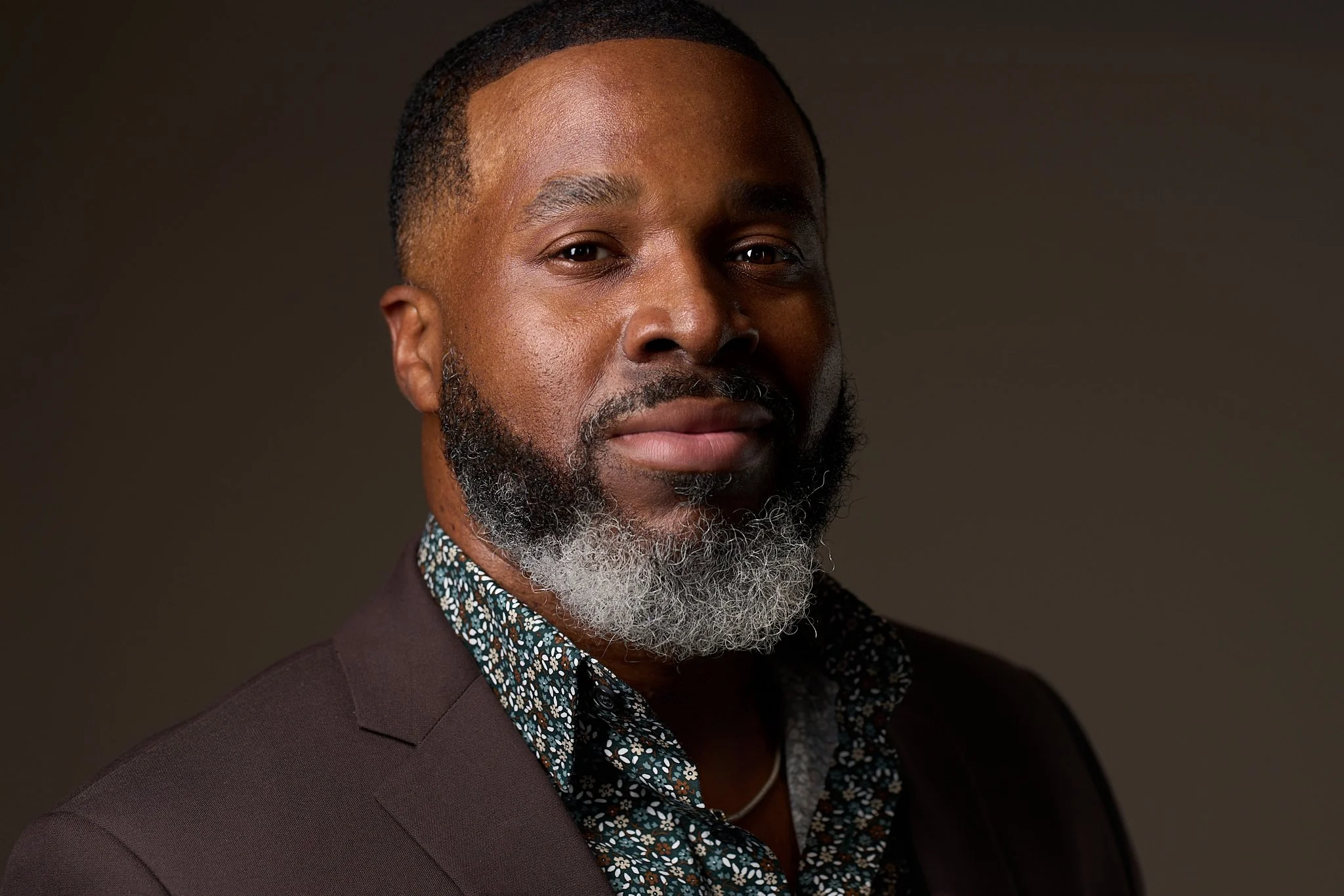 A portrait of an African American man with short hair and a long, grey beard, wearing a brown blazer and a patterned shirt, looking at the camera against a dark background.
