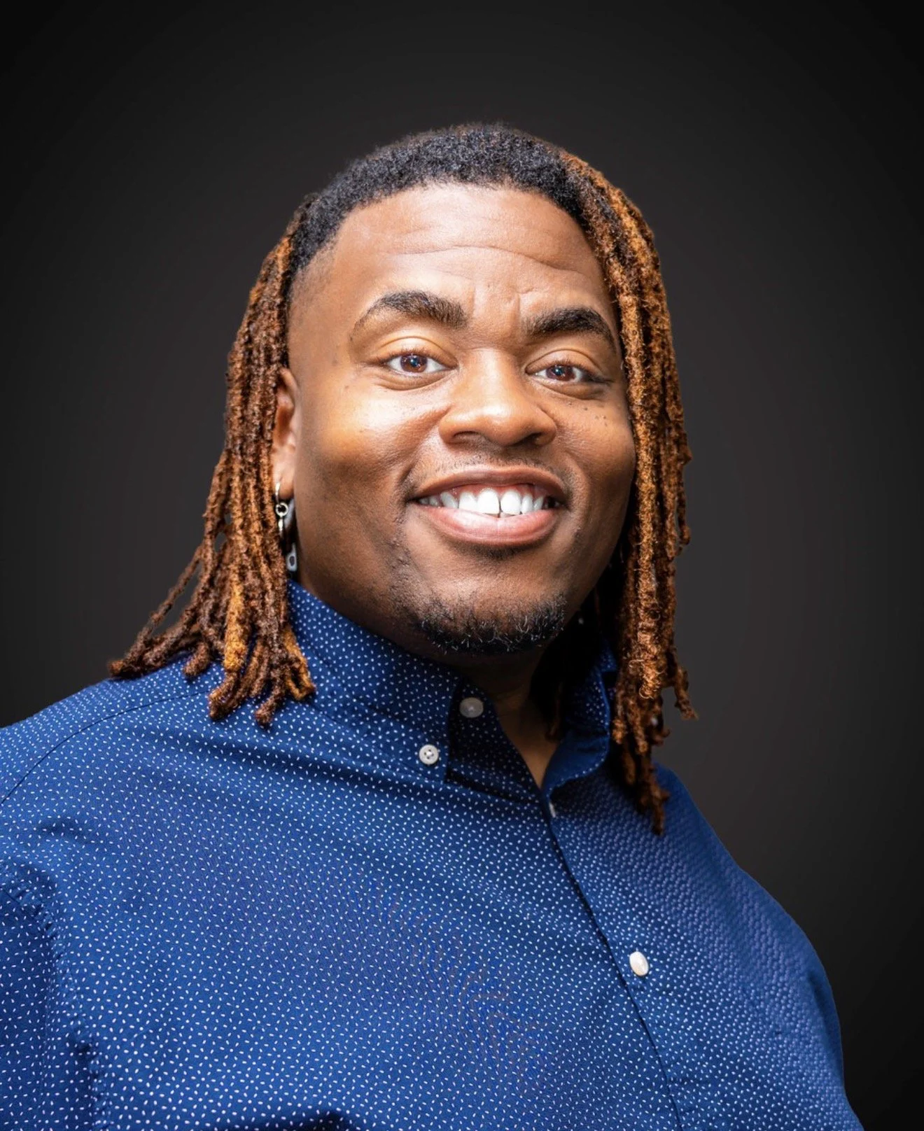 A smiling man with dreadlocks wearing a blue button-up shirt with small white dots against a dark background.