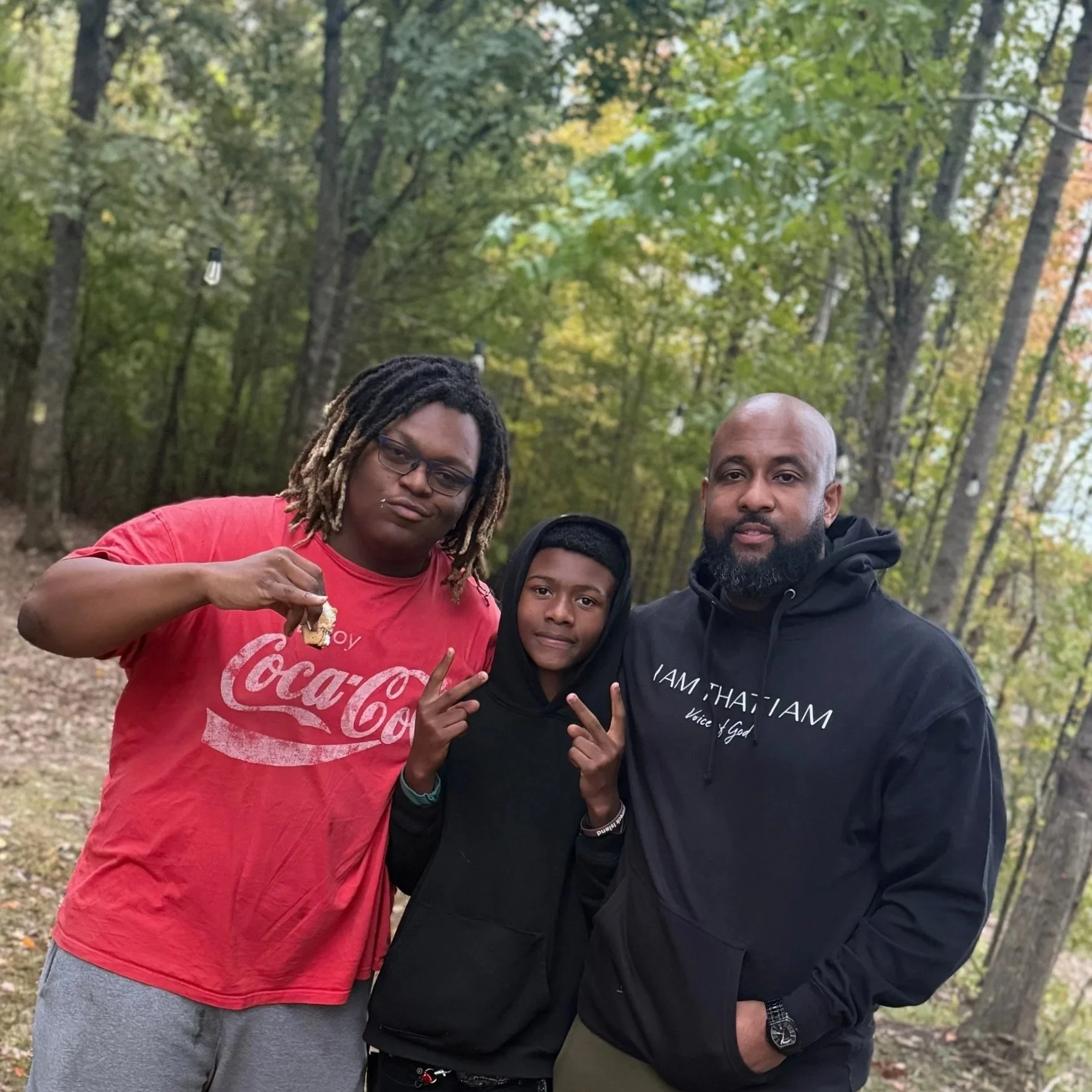 Three people posing outdoors in a wooded area, two men and a young boy. The man on the left has dreadlocks and glasses, wearing a red Coca-Cola t-shirt. The boy in the middle is making a peace sign with his hands, wearing a black hoodie. The man on the right has a beard, wearing a black hoodie with white text, and has his arm around the boy.