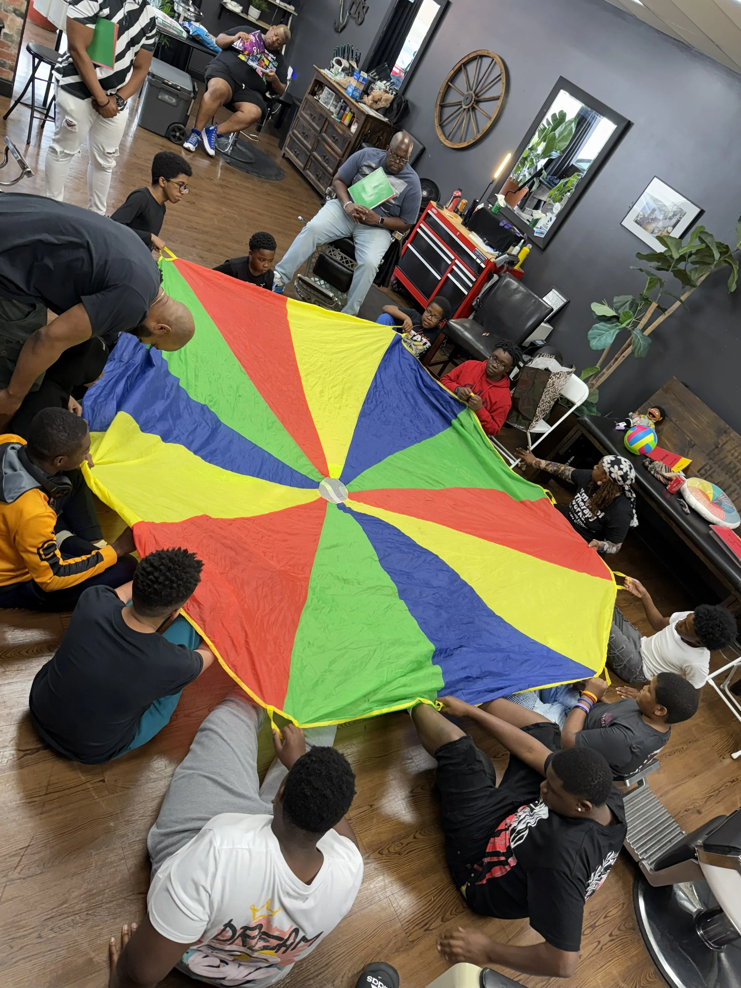 A group of people, including children and adults, gathered in a room playing with a colorful parachute. Some are sitting on the floor holding the parachute, while others are standing around. The room has wooden floors and dark-colored walls with various decorations, plants, and furniture.