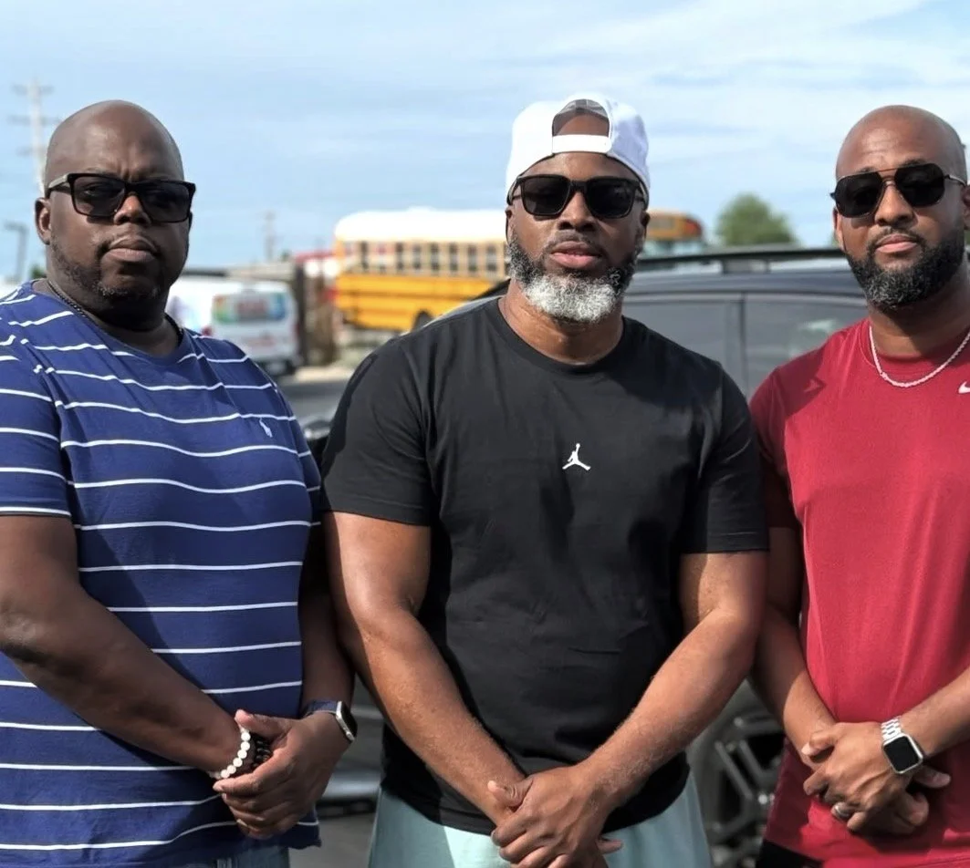 Three men wearing sunglasses and casual clothing standing outdoors with a bus and vehicles in background.