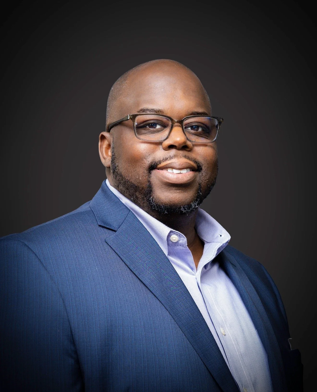 A portrait of a man with glasses, dressed in a blue suit and white shirt, smiling at the camera against a dark background.