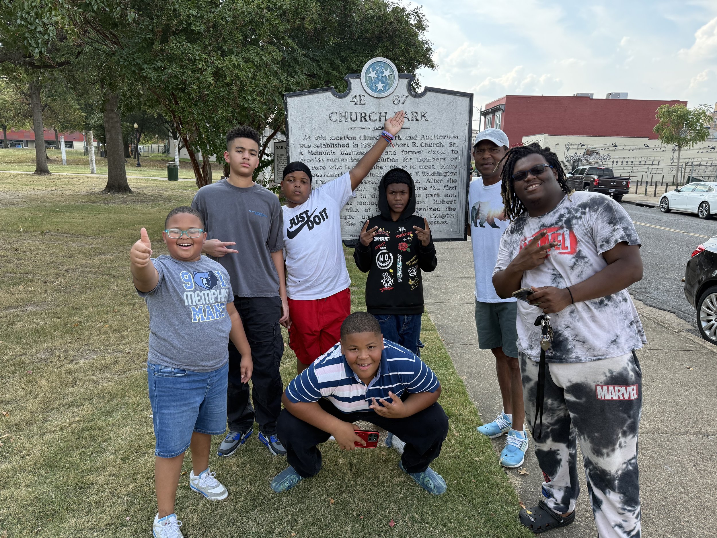 A group of seven people, including children and adults, are standing on a sidewalk in front of a historical marker about a church. They are smiling, making gestures, and posing for the photo on a grassy area with trees and parking lots in the background.